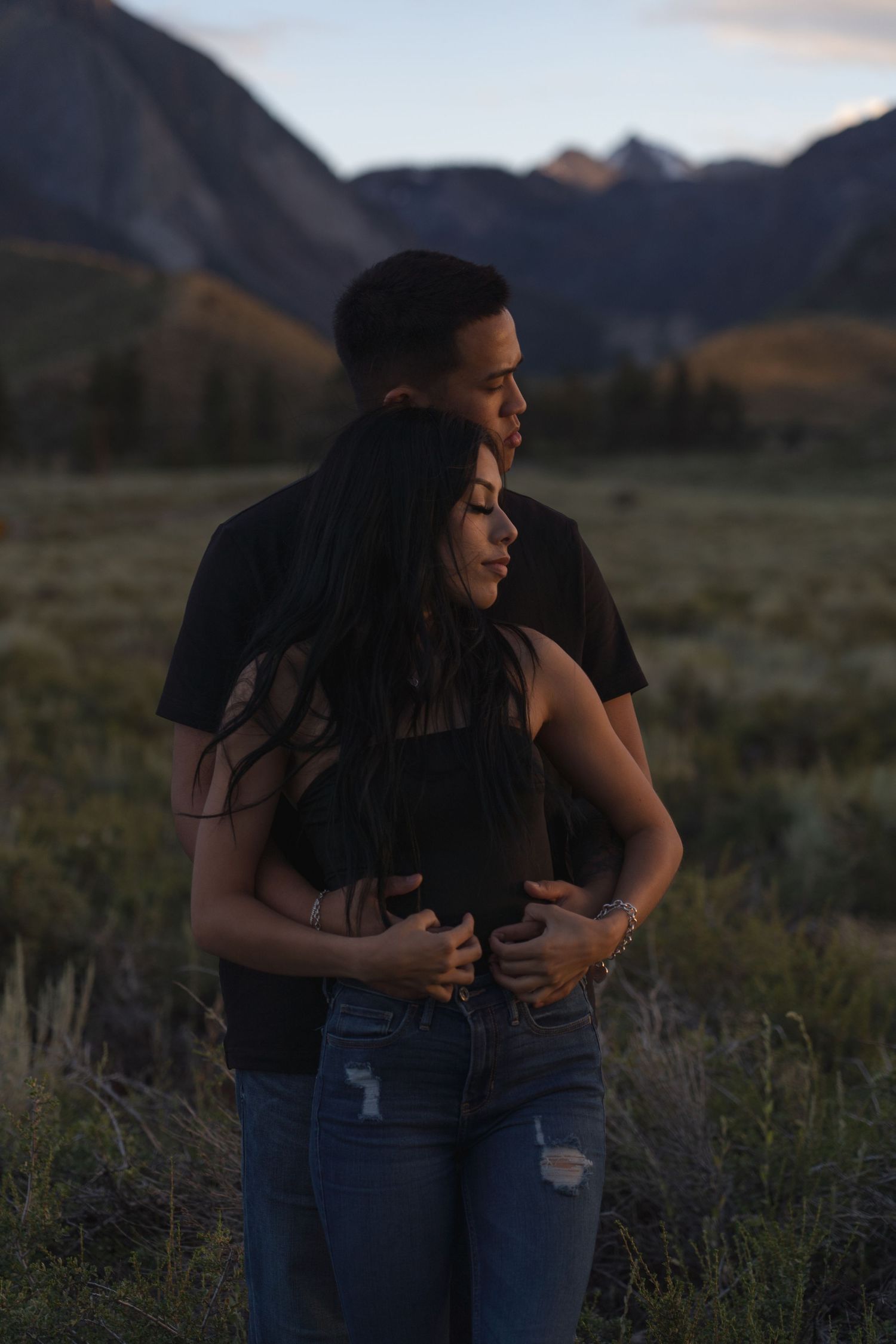 A couple embraces in a scenic mountain landscape during golden hour in casual attire.