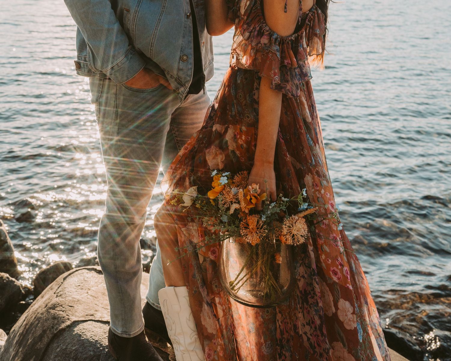 A couple stands on rocky coastline during sunset while ocean waves sparkle in the background.