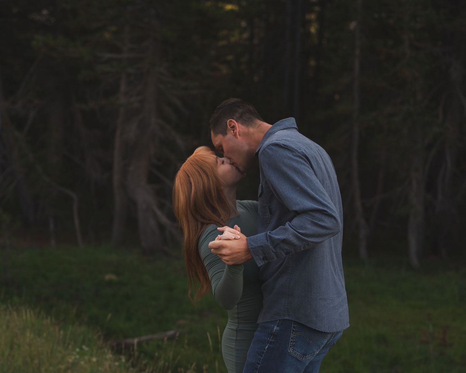 A couple shares a romantic kiss while embracing outdoors in a lush green forest setting at dusk.
