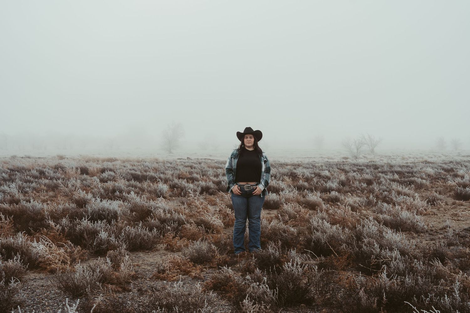 Person wearing a hat and winter clothing stands in a misty field of dried sagebrush on an overcast day.