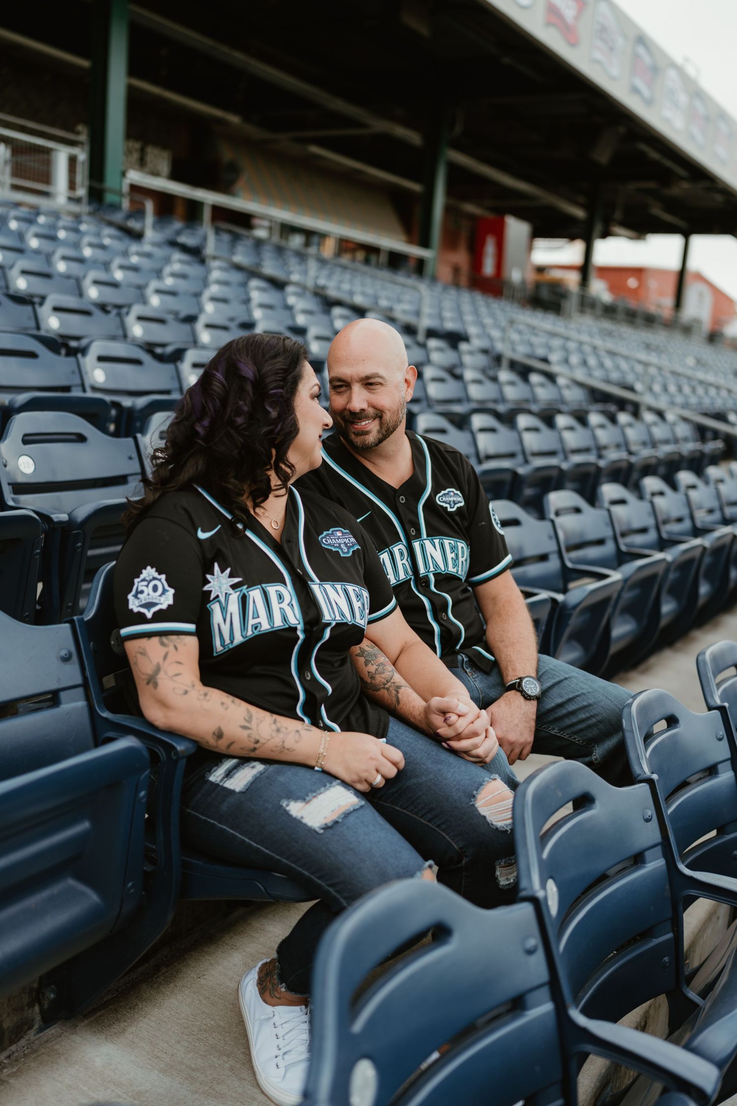 Couple in matching Seattle Mariners jerseys sitting together in stadium seats, smiling at each other.
