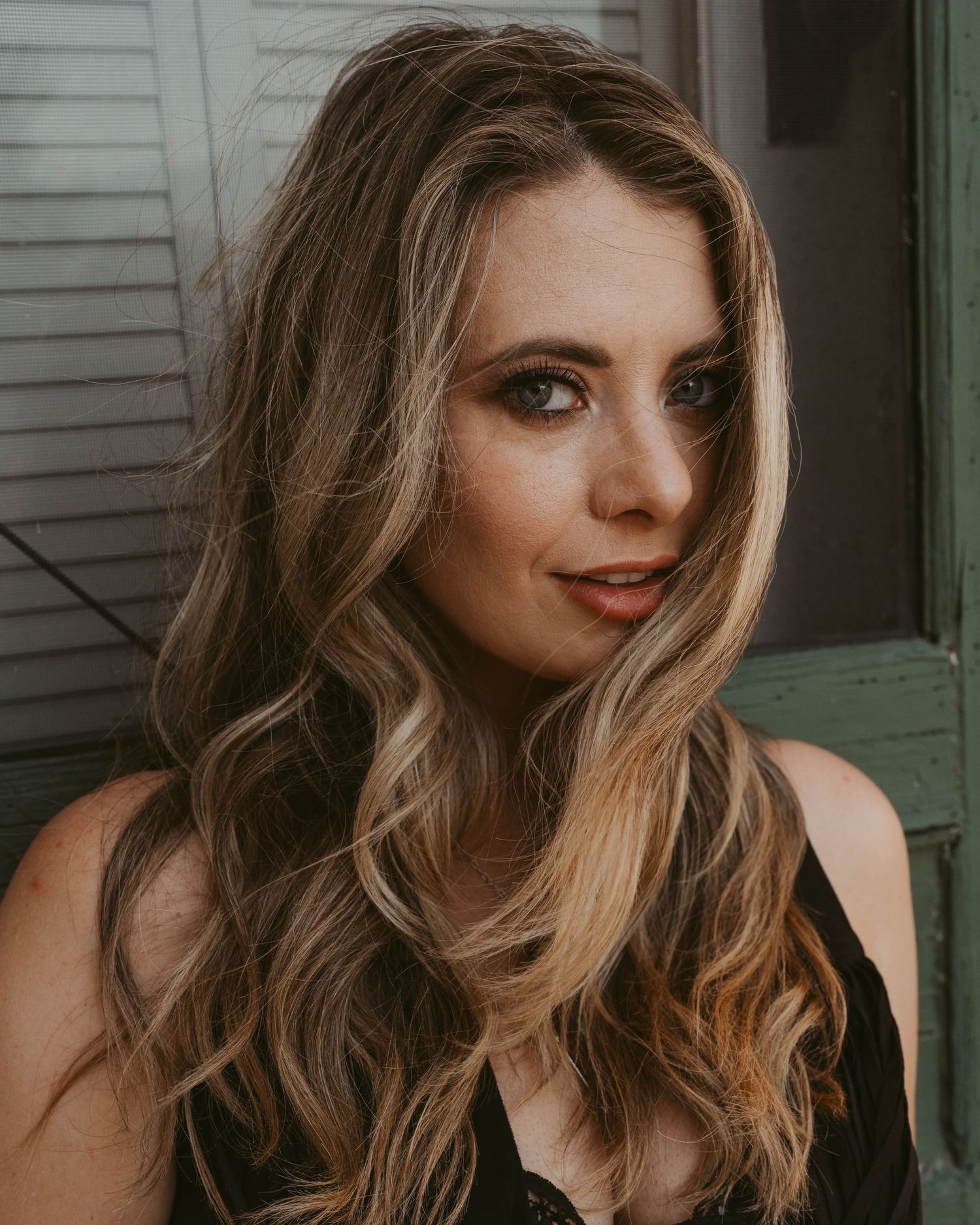 Portrait with blonde wavy hair wearing moody makeup against white siding in warm natural lighting.