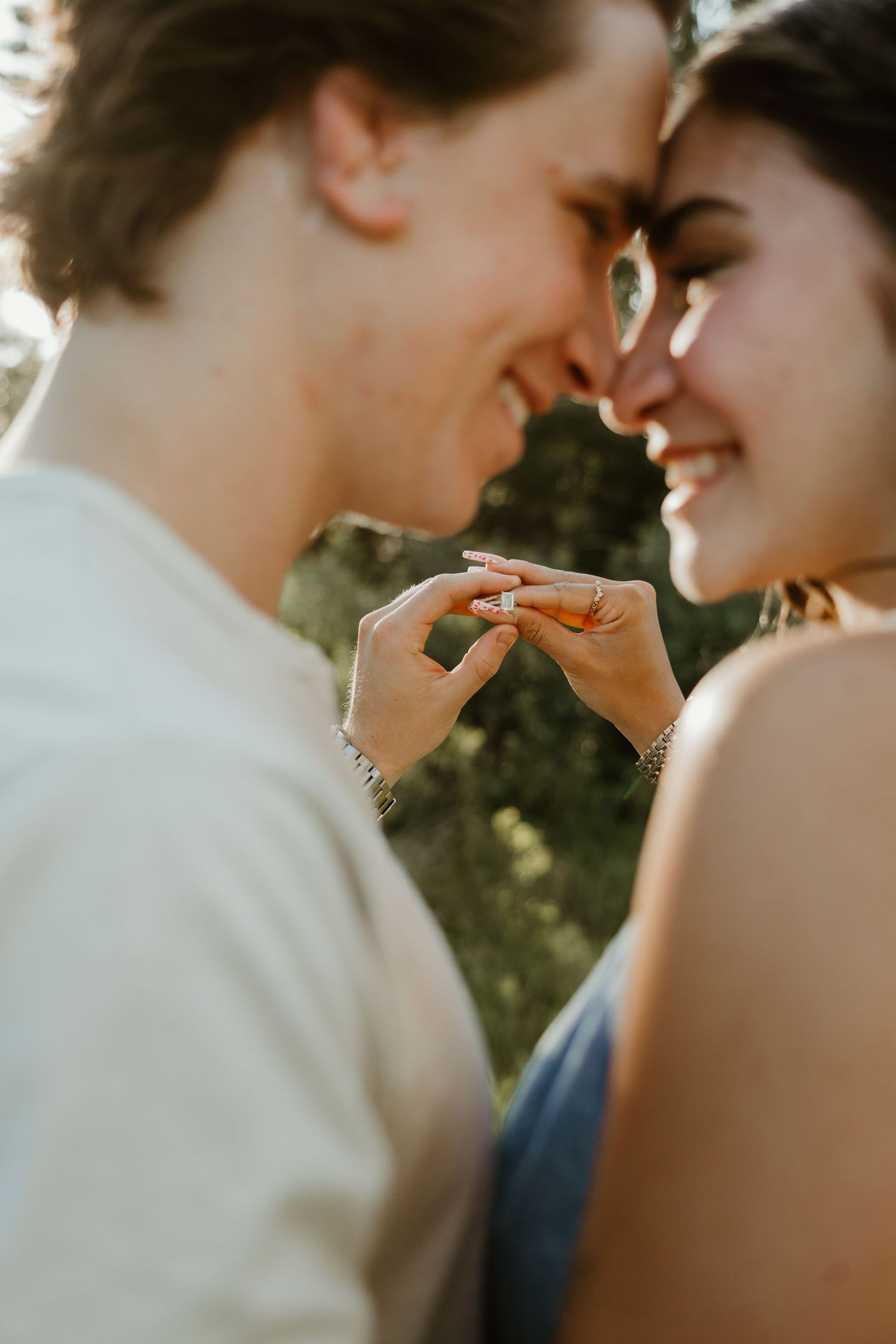 A smiling couple touches pinkies romantically while leaning foreheads together outdoors in warm sunlight.