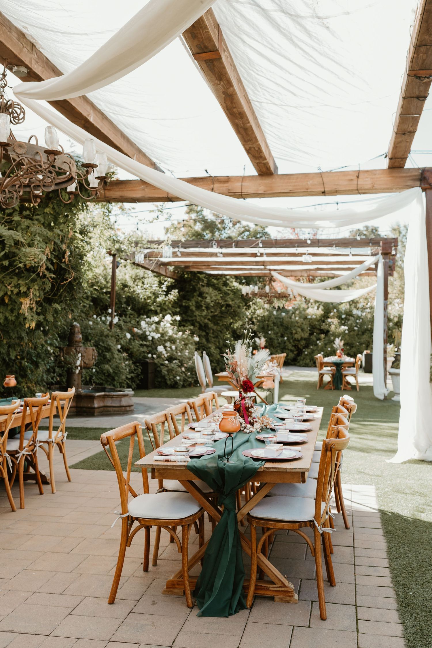 Outdoor wedding reception setup with wooden tables and chairs under white draped canopy and green floral decor.