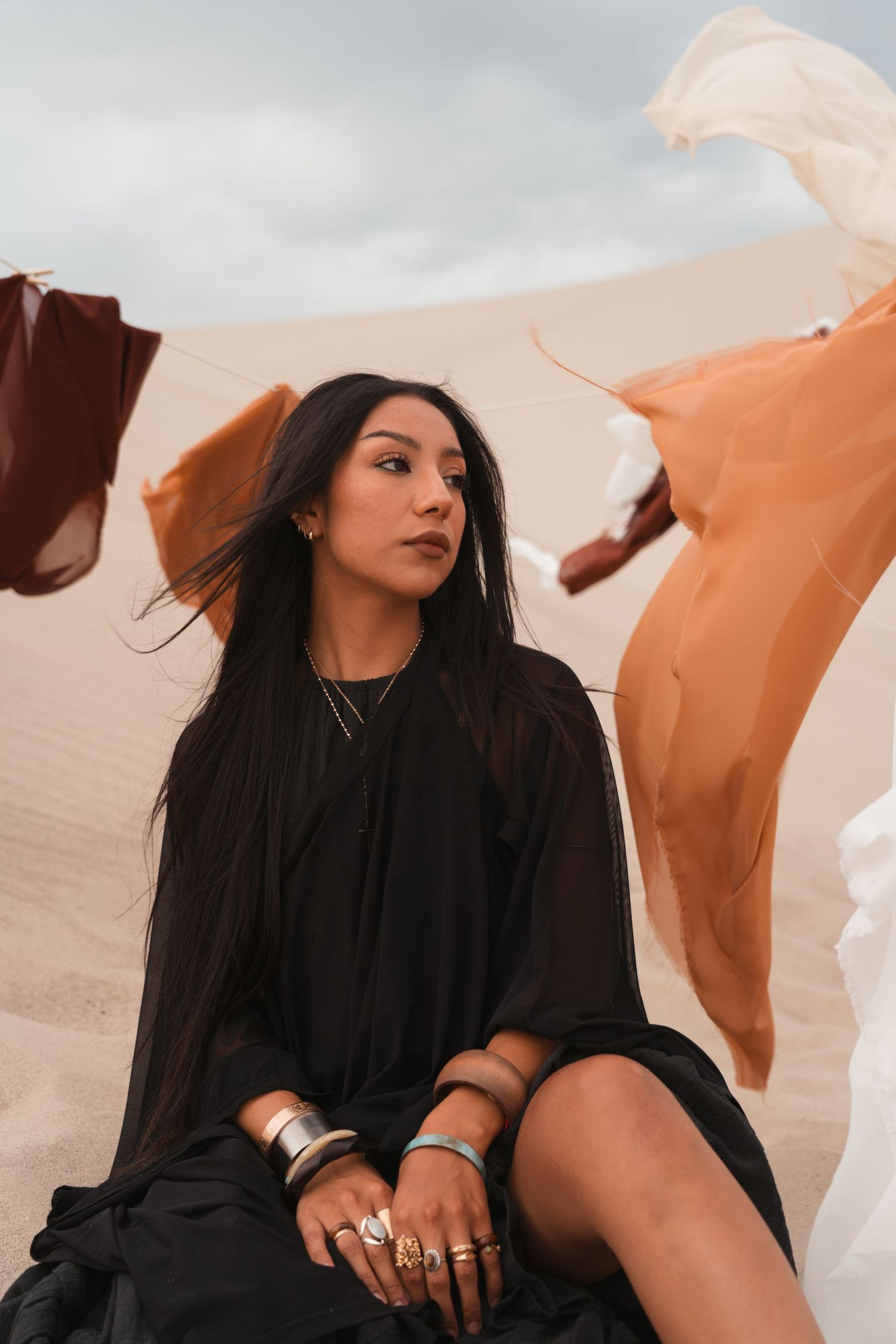 Woman in black dress sitting in desert sand dunes, adorned with silver jewelry, colorful fabric flowing in wind.