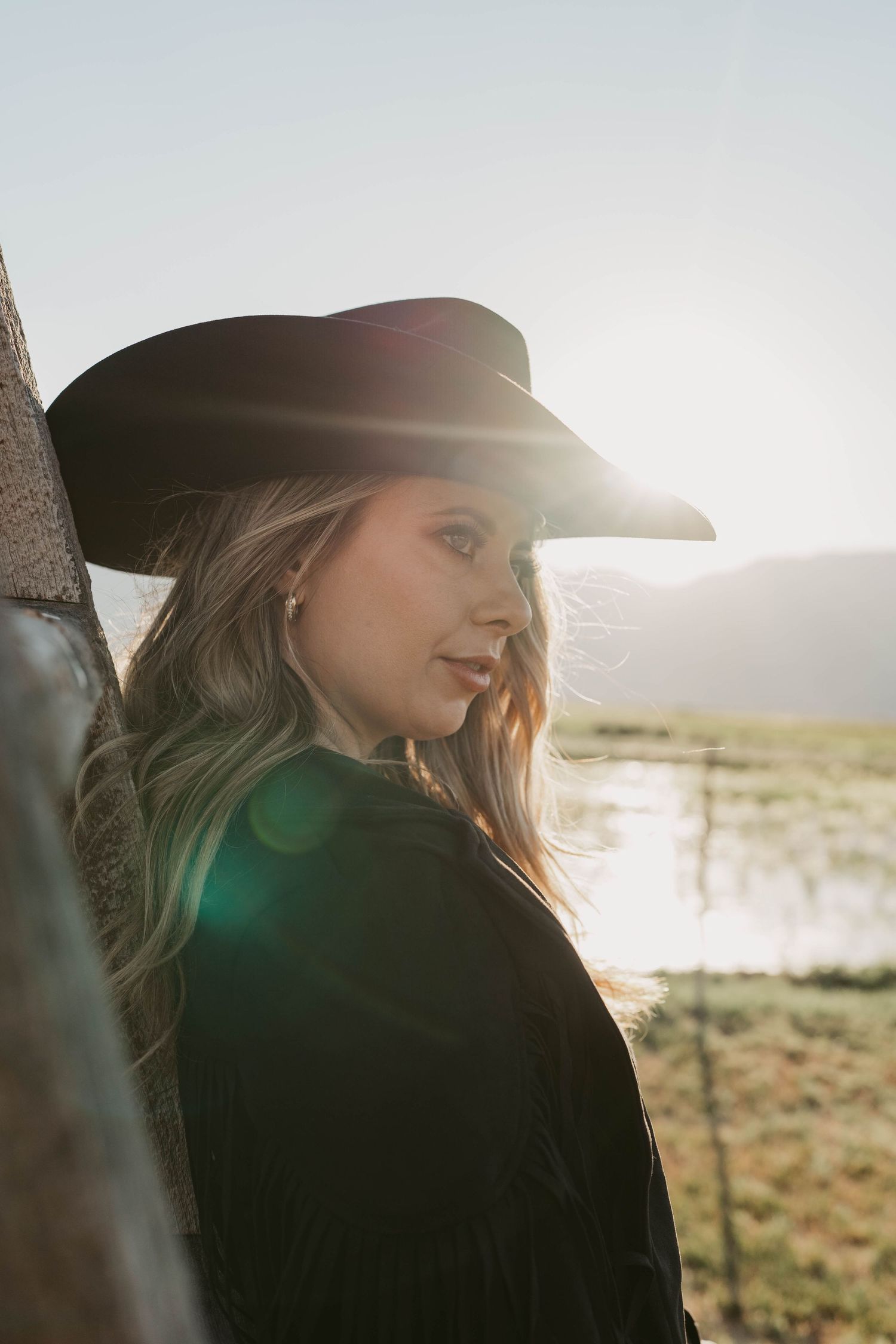A person in a black cowboy hat and jacket poses outdoors during golden hour with sunlight creating a backlit silhouette.