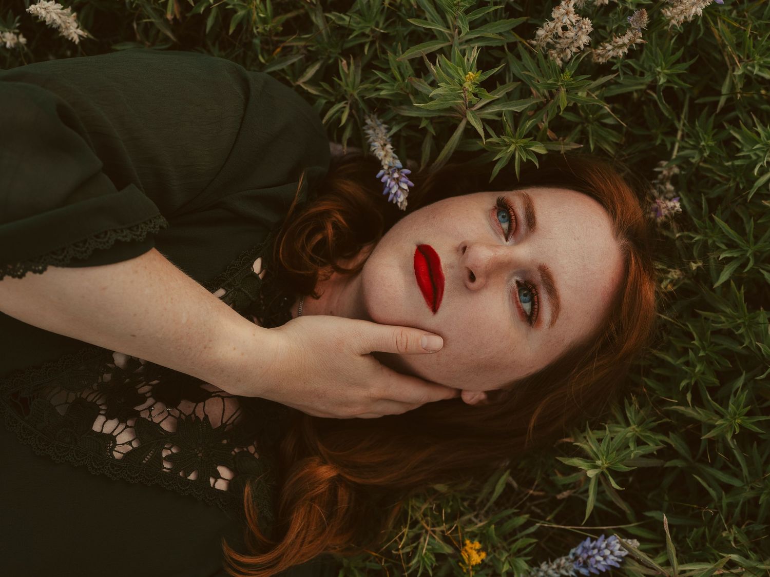 A young woman with red hair and bold red lips lies among green plants and wildflowers in a dreamy outdoor portrait.