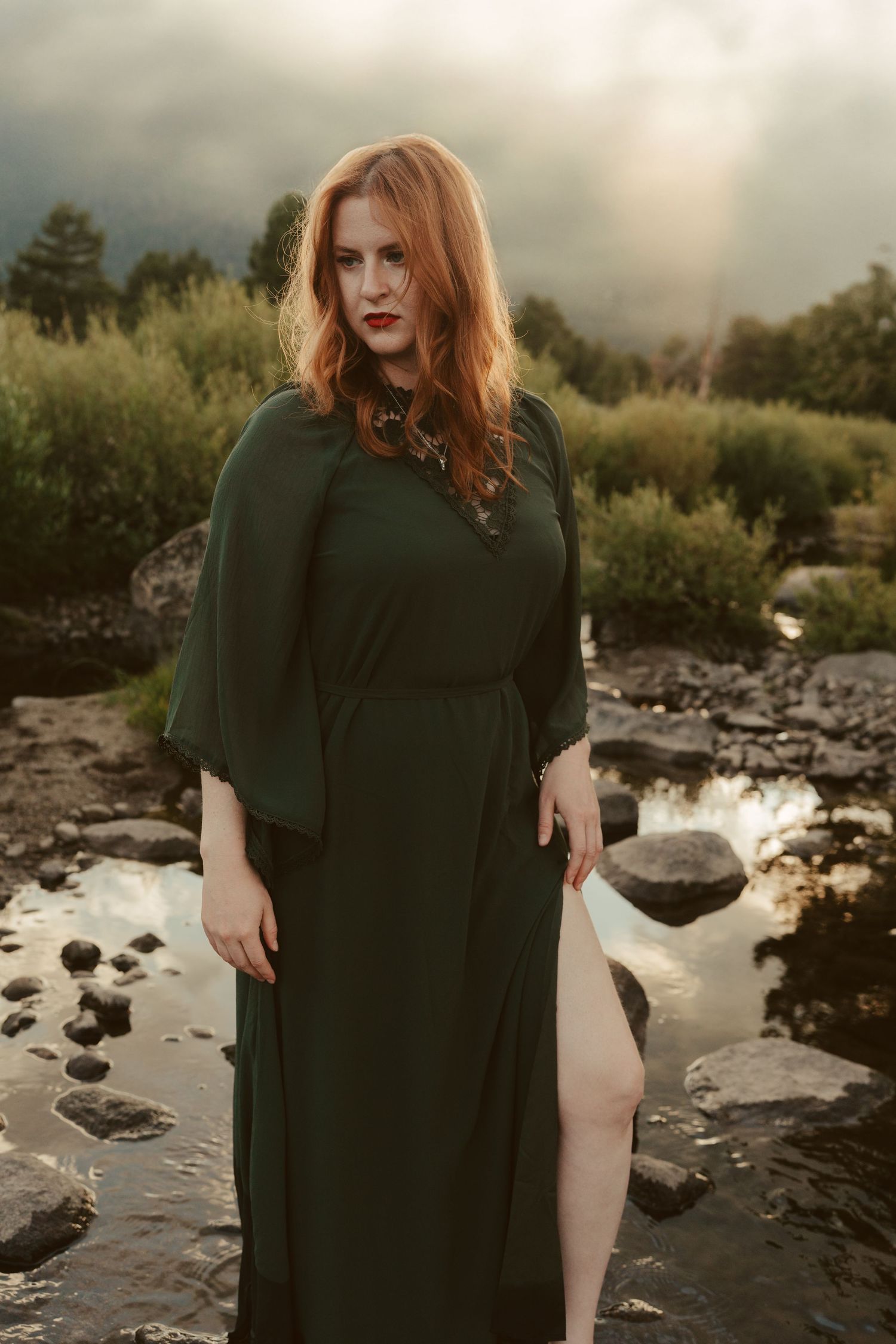 A woman in a dark green dress poses near a rocky riverbank during sunset.
