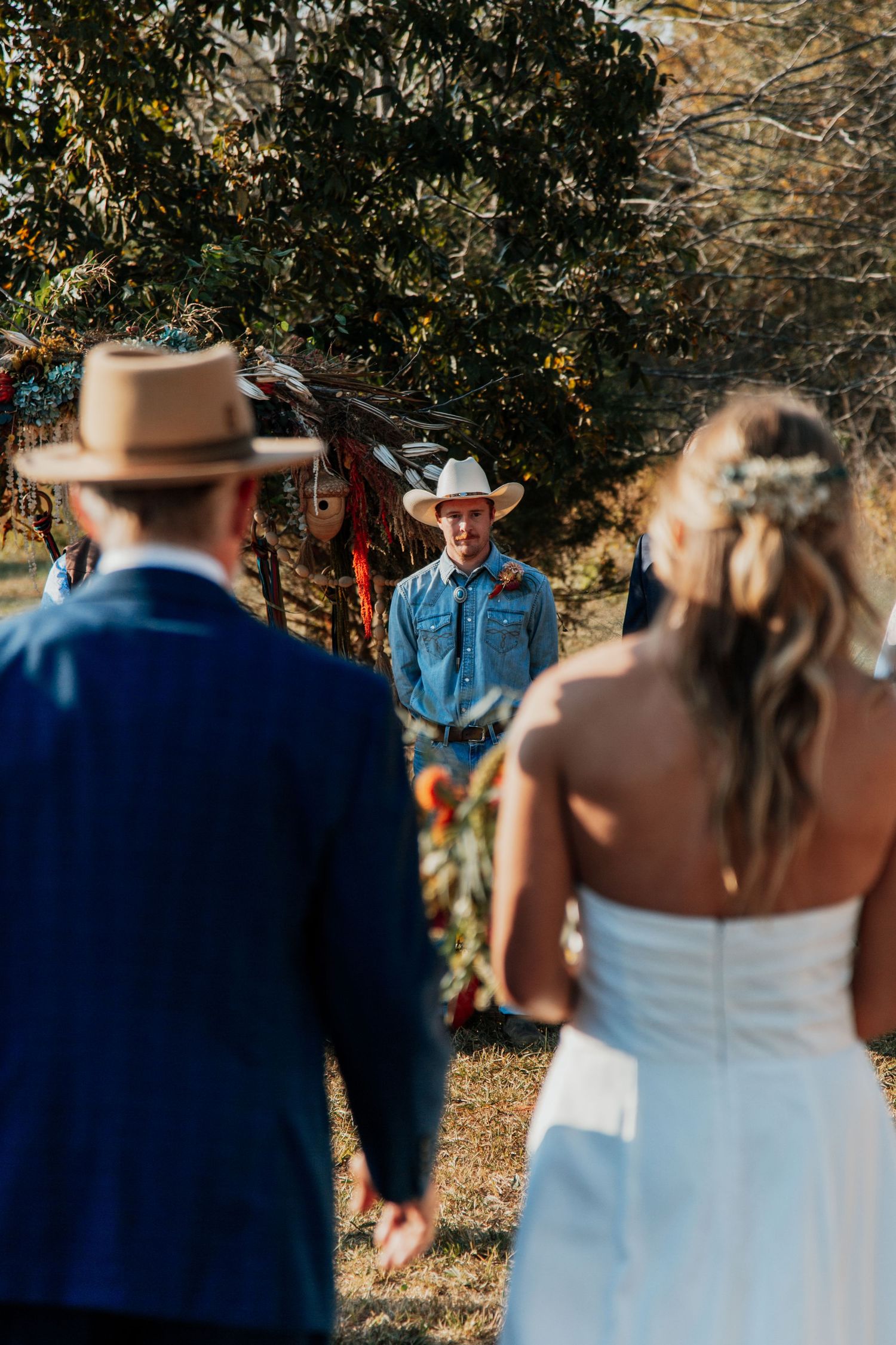 Outdoor rustic wedding ceremony with bride and groom holding hands facing officiant in cowboy hat near floral arch.