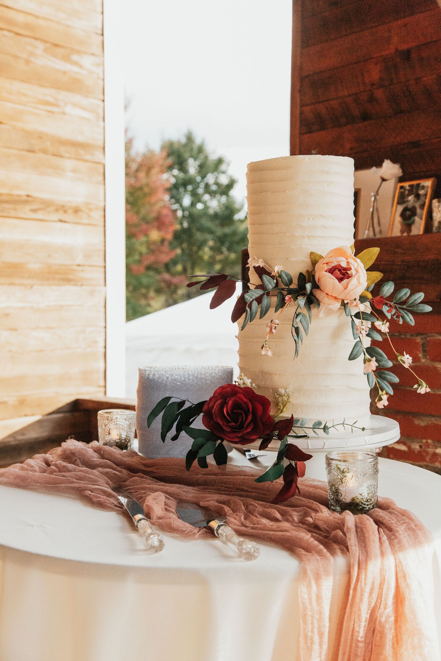 Two-tier white wedding cake adorned with blush peonies, burgundy roses, and greenery on a draped table.