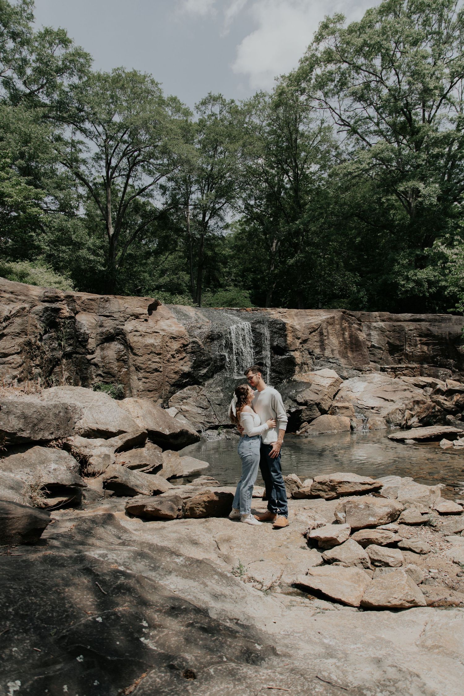 Couple embracing on rocky terrain beside a serene waterfall surrounded by lush green trees in a natural park setting.