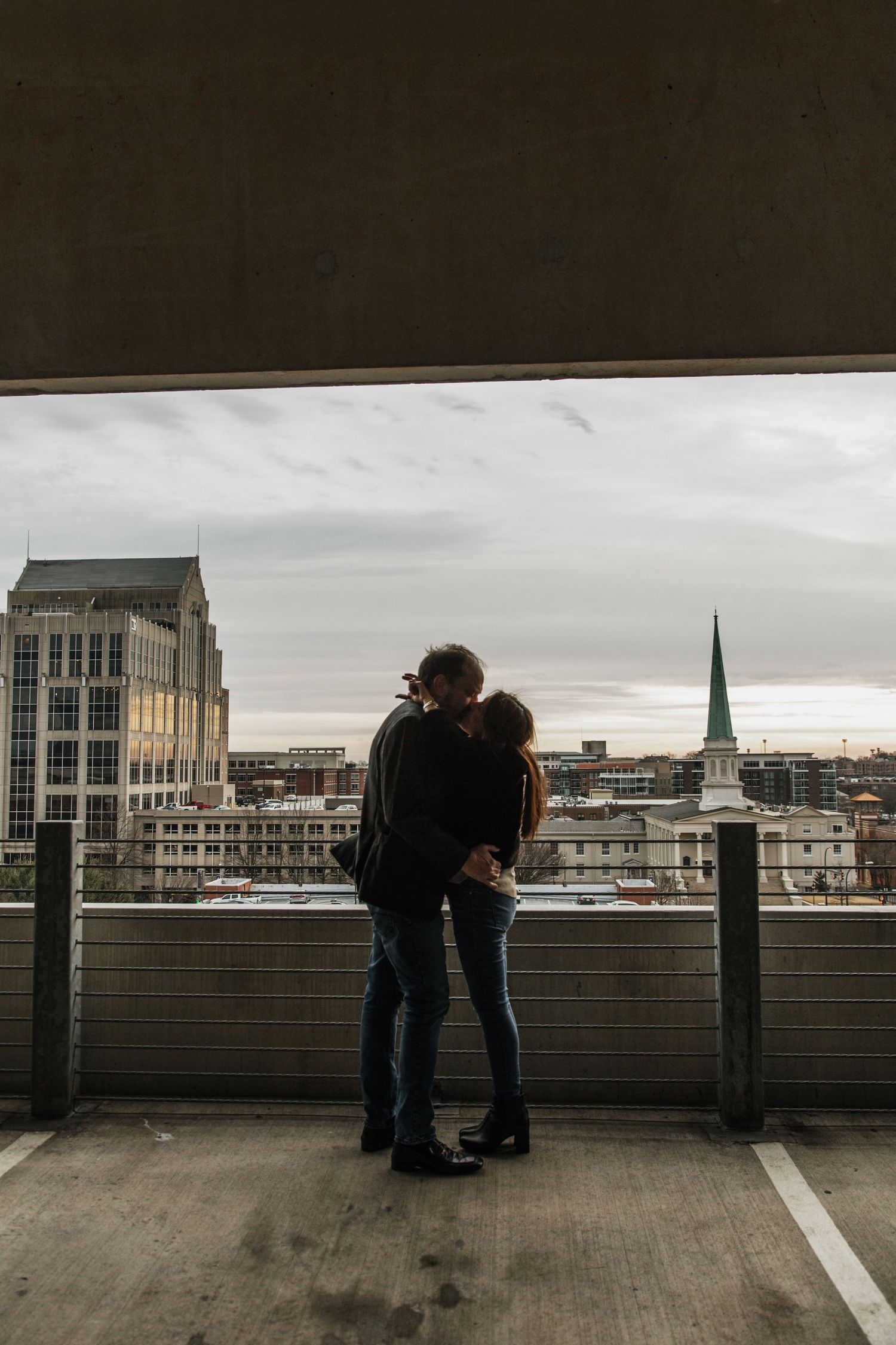 Couple embracing on urban rooftop parking garage overlooking city skyline with church steeple on cloudy day.