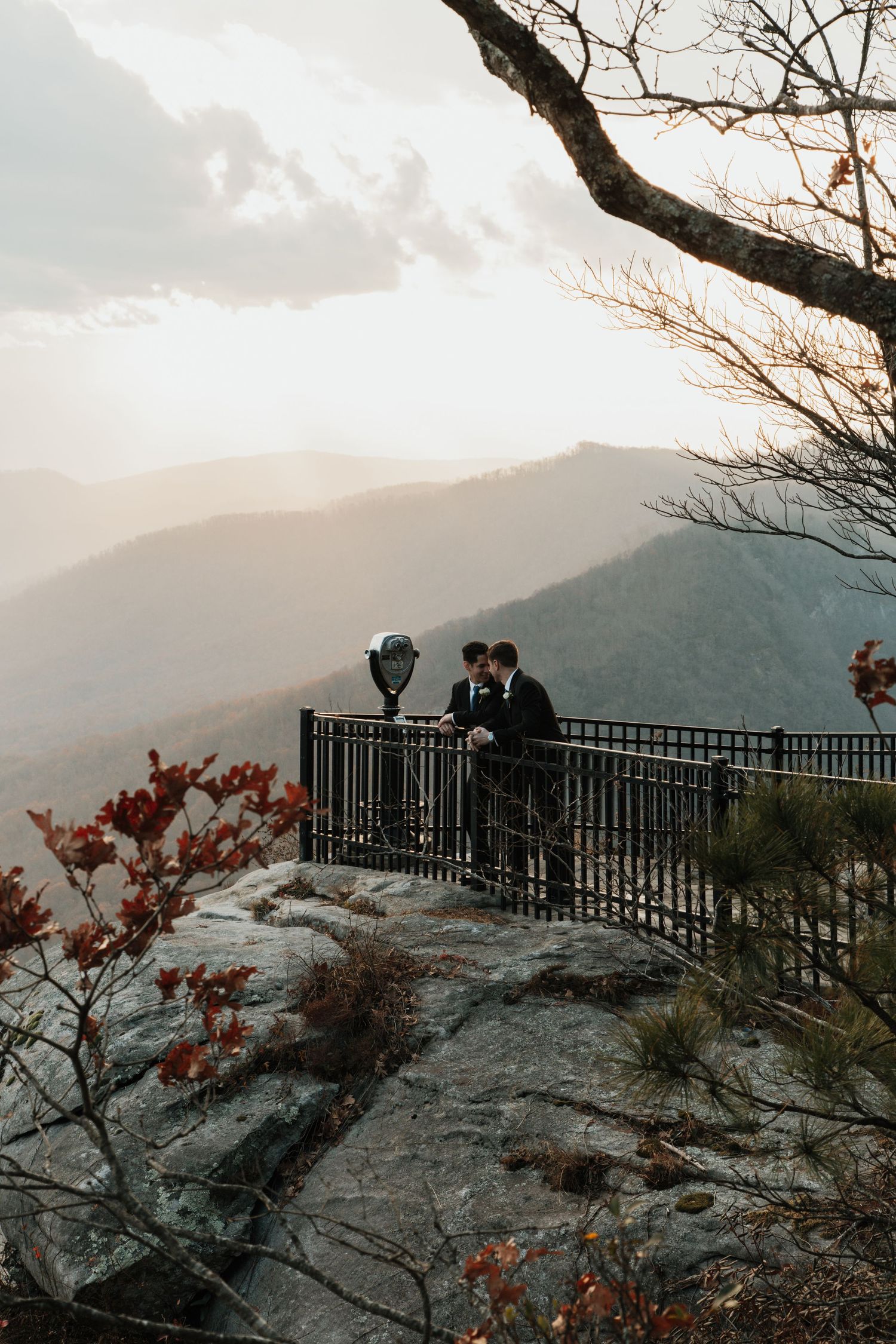 Two grooms share a romantic moment at a mountain overlook with scenic valley views at sunset.