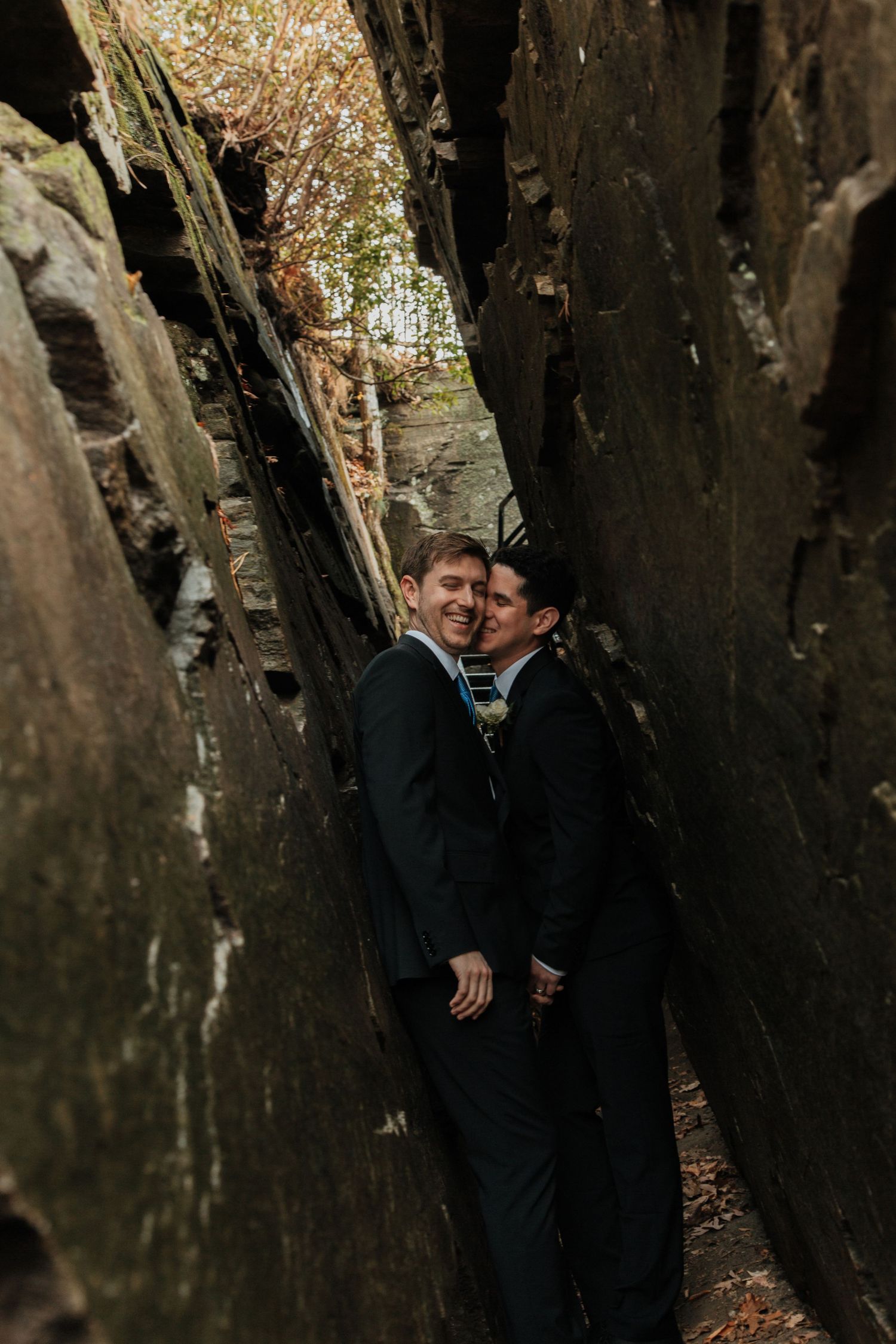 Two grooms in dark suits share a tender moment between towering rock formations in a forest setting.