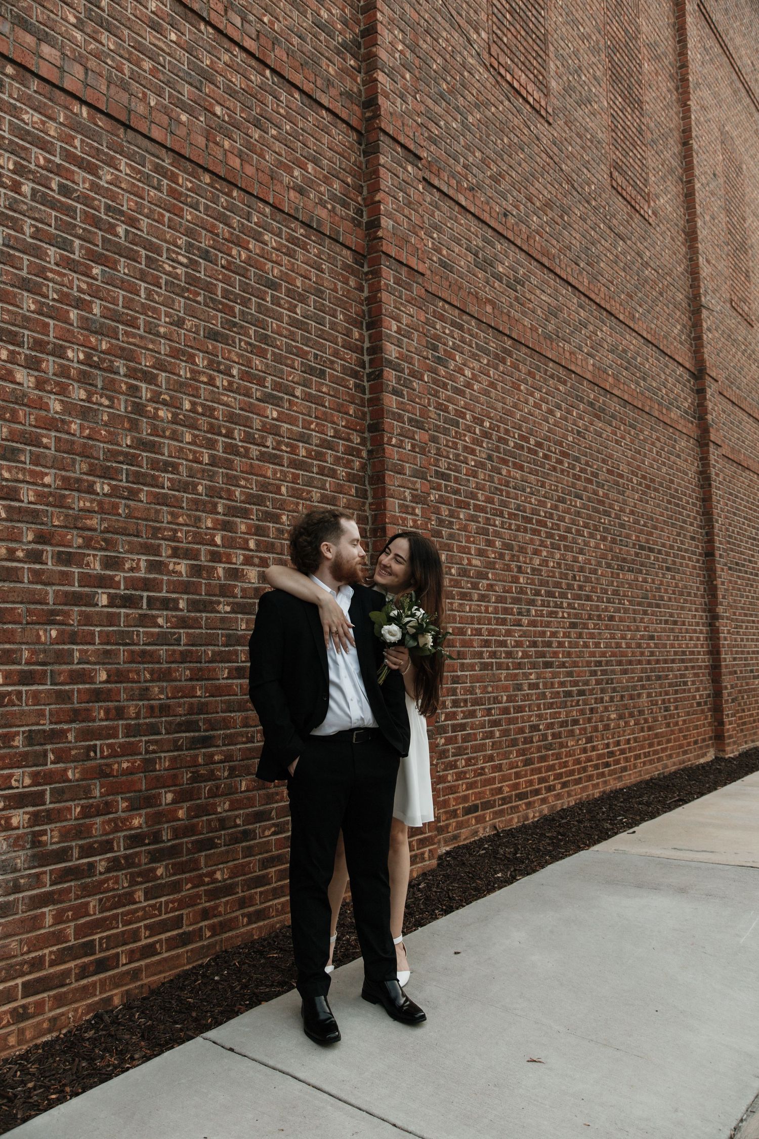 Wedding couple embracing against rustic brick wall, bride in short white dress holding bouquet.