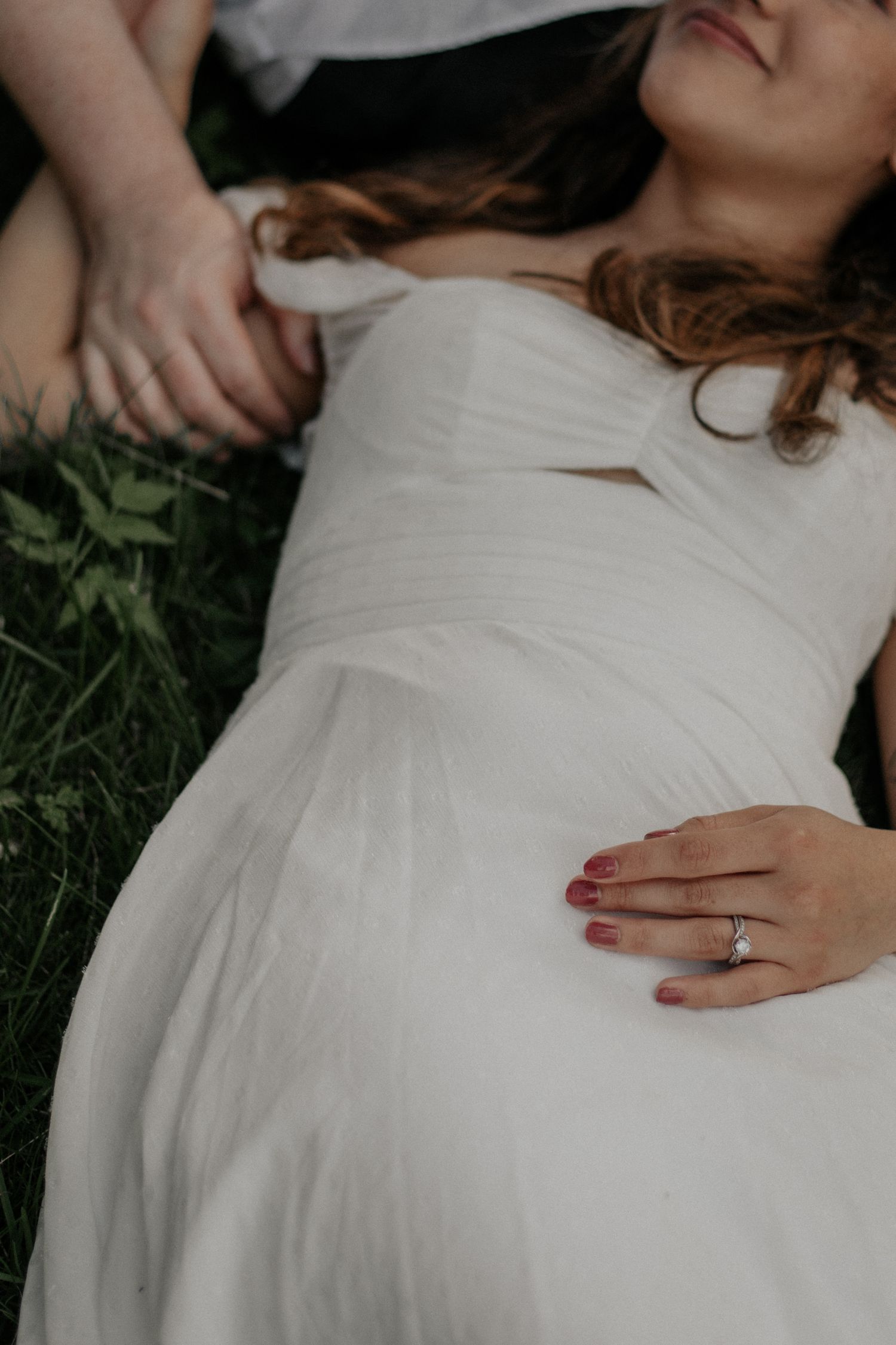 Bride in white gown lying in grass displaying her engagement ring.