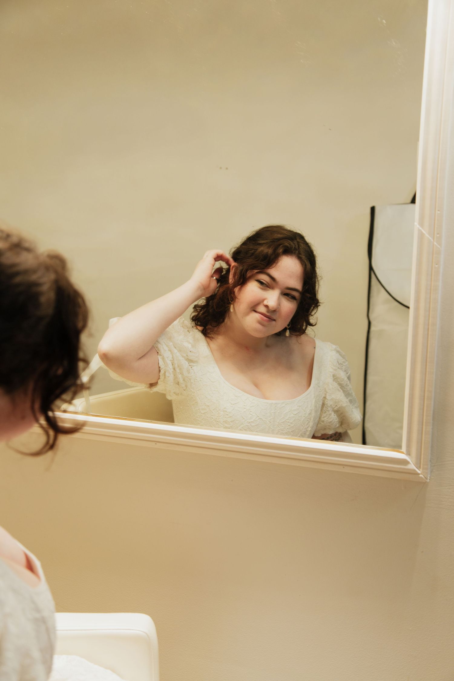 Bride in white lace dress smiling at reflection while styling curly hair in bathroom mirror. venue at ballenger bridge landrum sc
