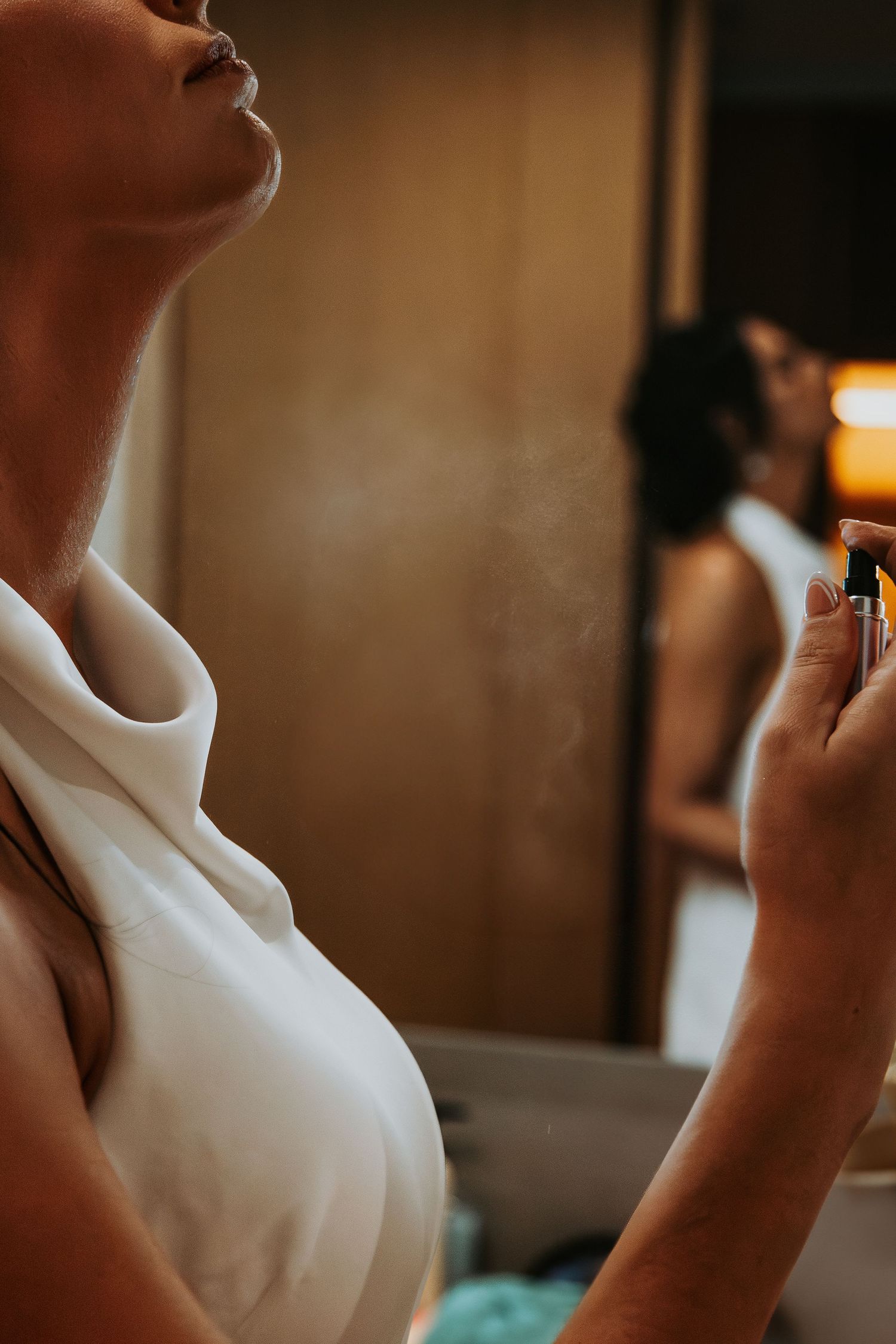 Bride in white robe sprays perfume on neck while getting ready in mirror. - riviera maya resort (cancun, Mexico)
