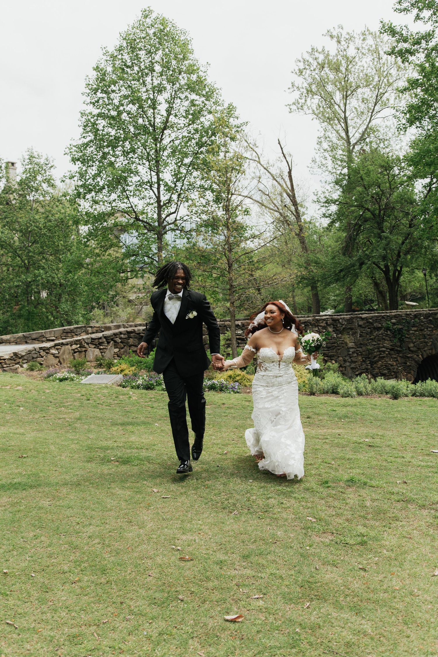 A joyful bride and groom in wedding attire run hand-in-hand across a lush green lawn surrounded by trees.