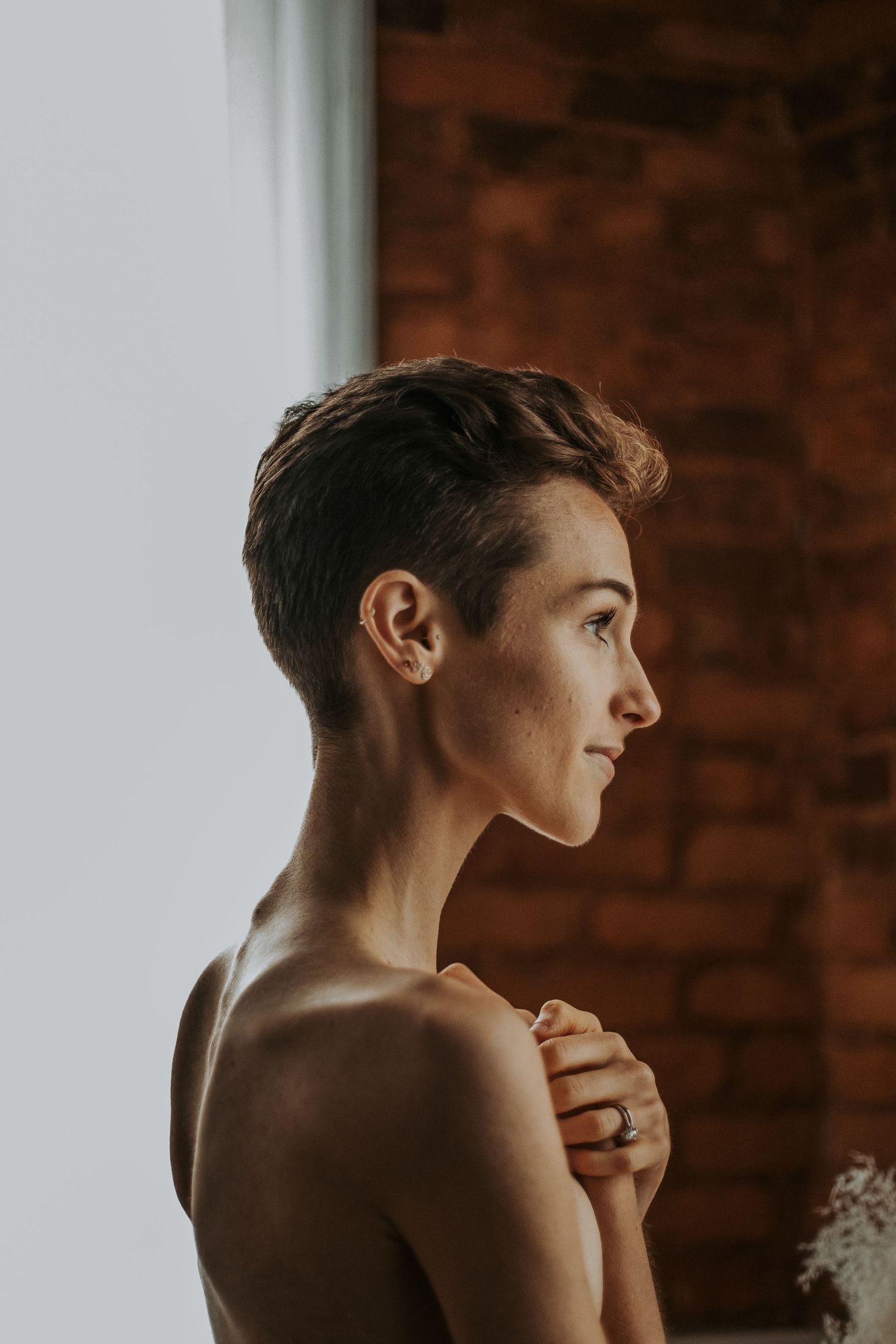 A woman with short hair gazes thoughtfully toward a window, hands clasped at her chest in soft natural light.