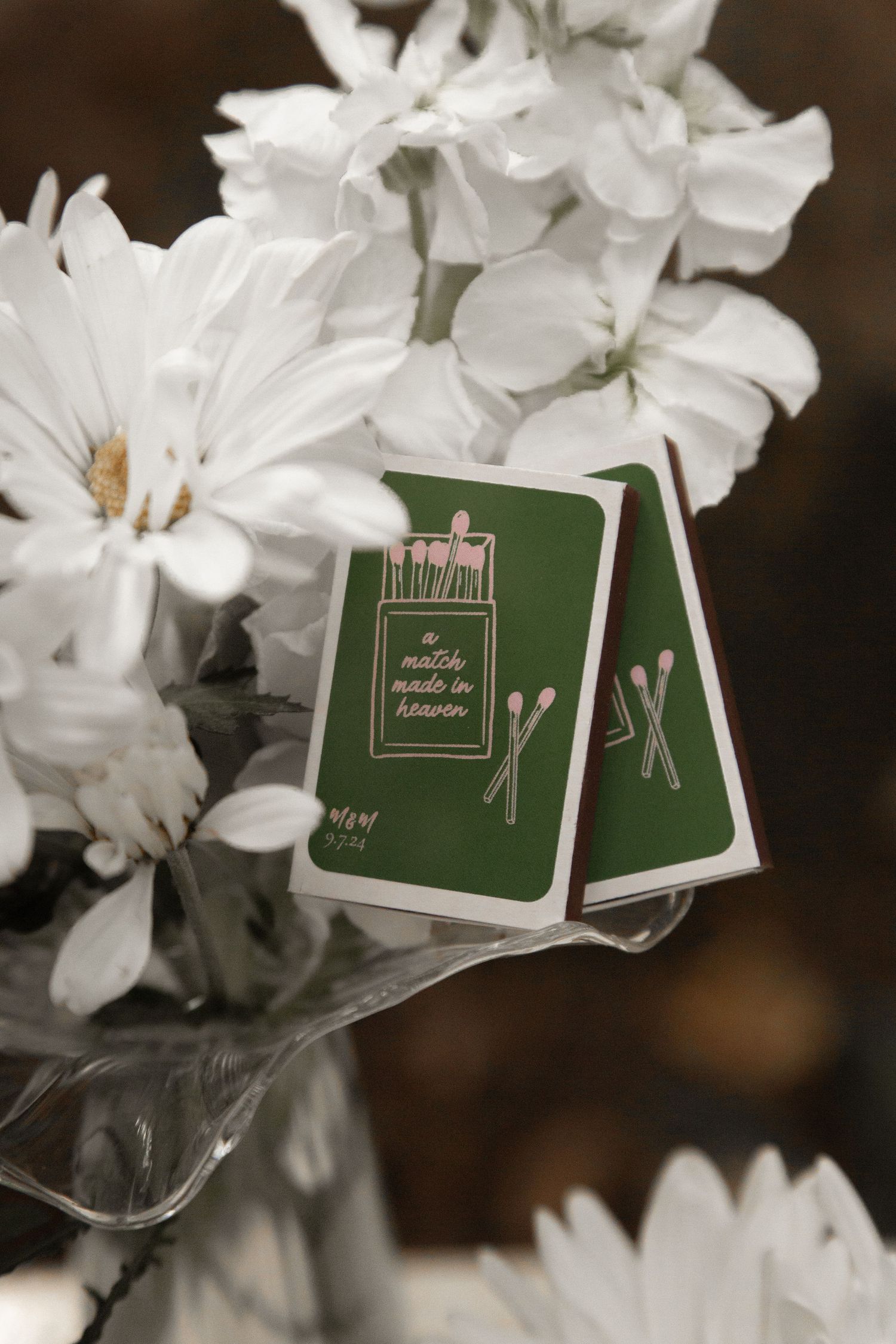 Green matchbook wedding favors reading 'a match made in heaven' nestled among white flowers in a glass vase.