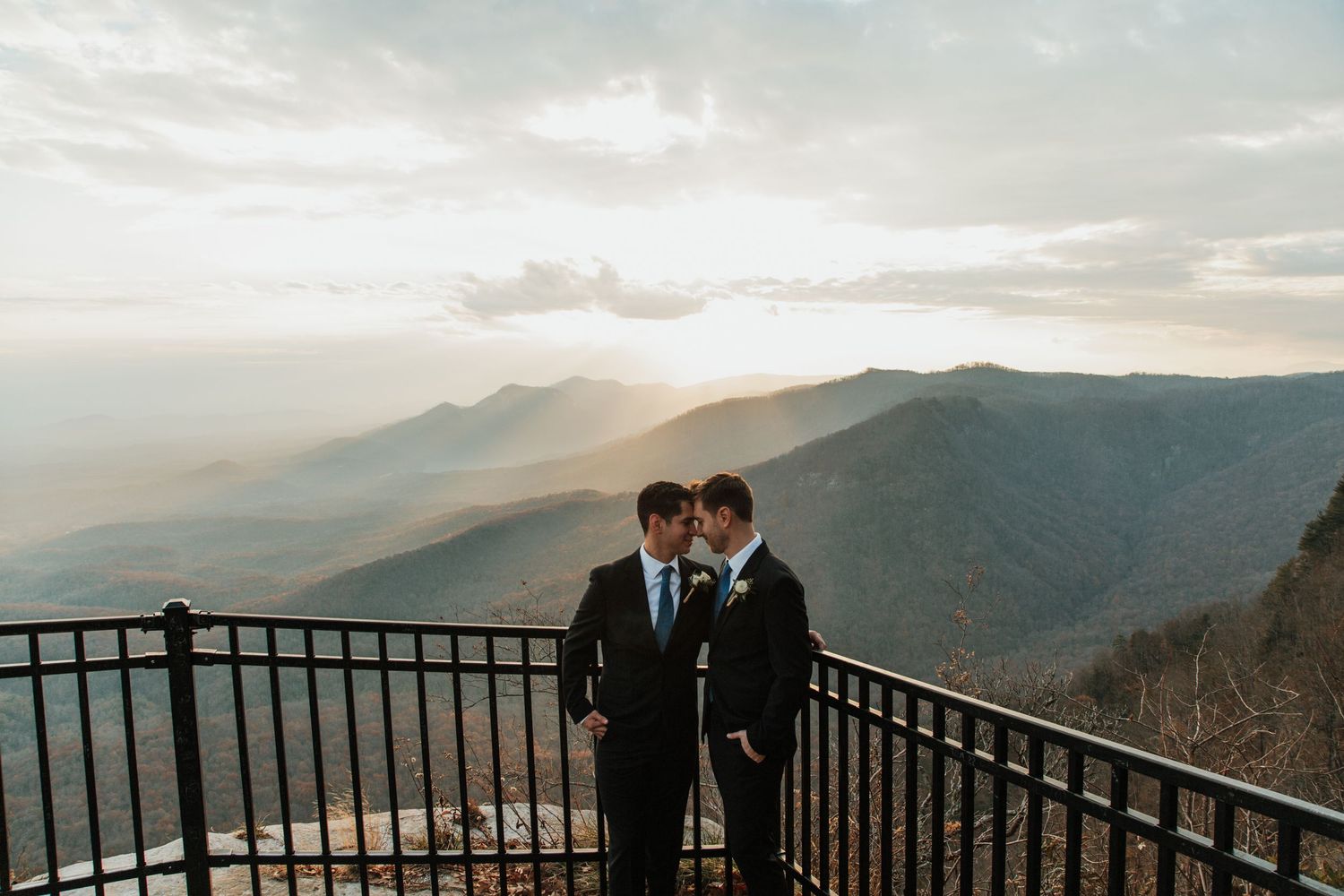 Two grooms share an intimate moment on a mountain overlook at sunset during their wedding day.