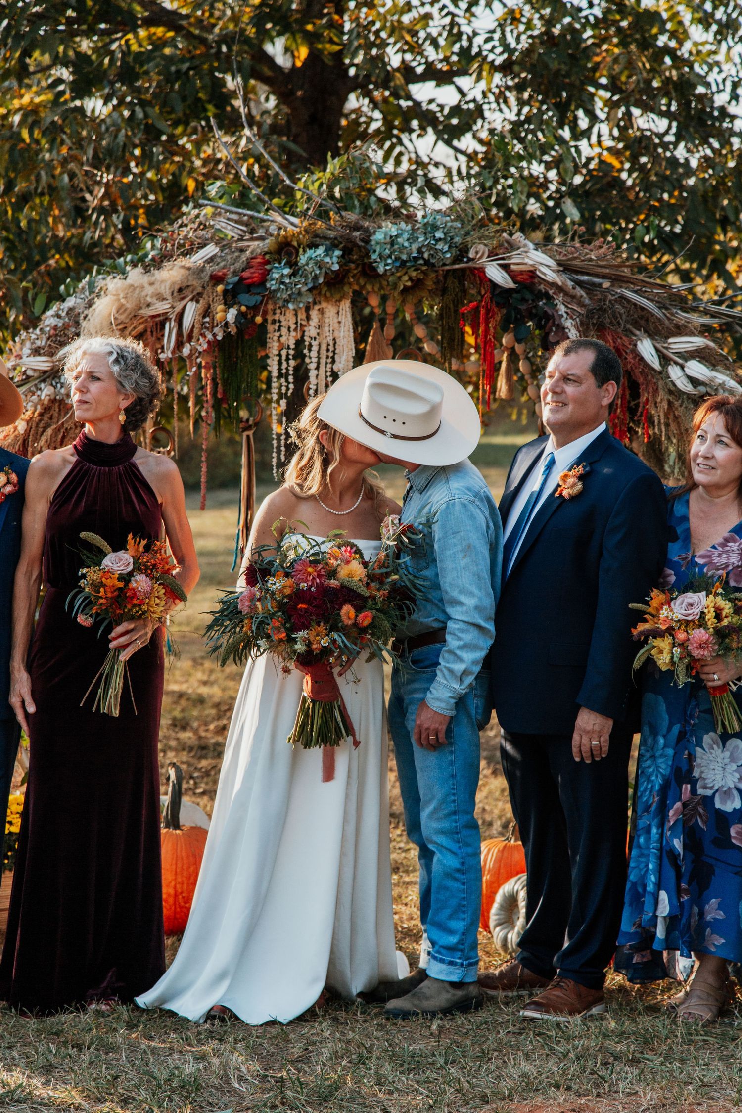 Bride and groom share a kiss at outdoor fall wedding ceremony with boho floral arch and autumn pumpkin decorations.