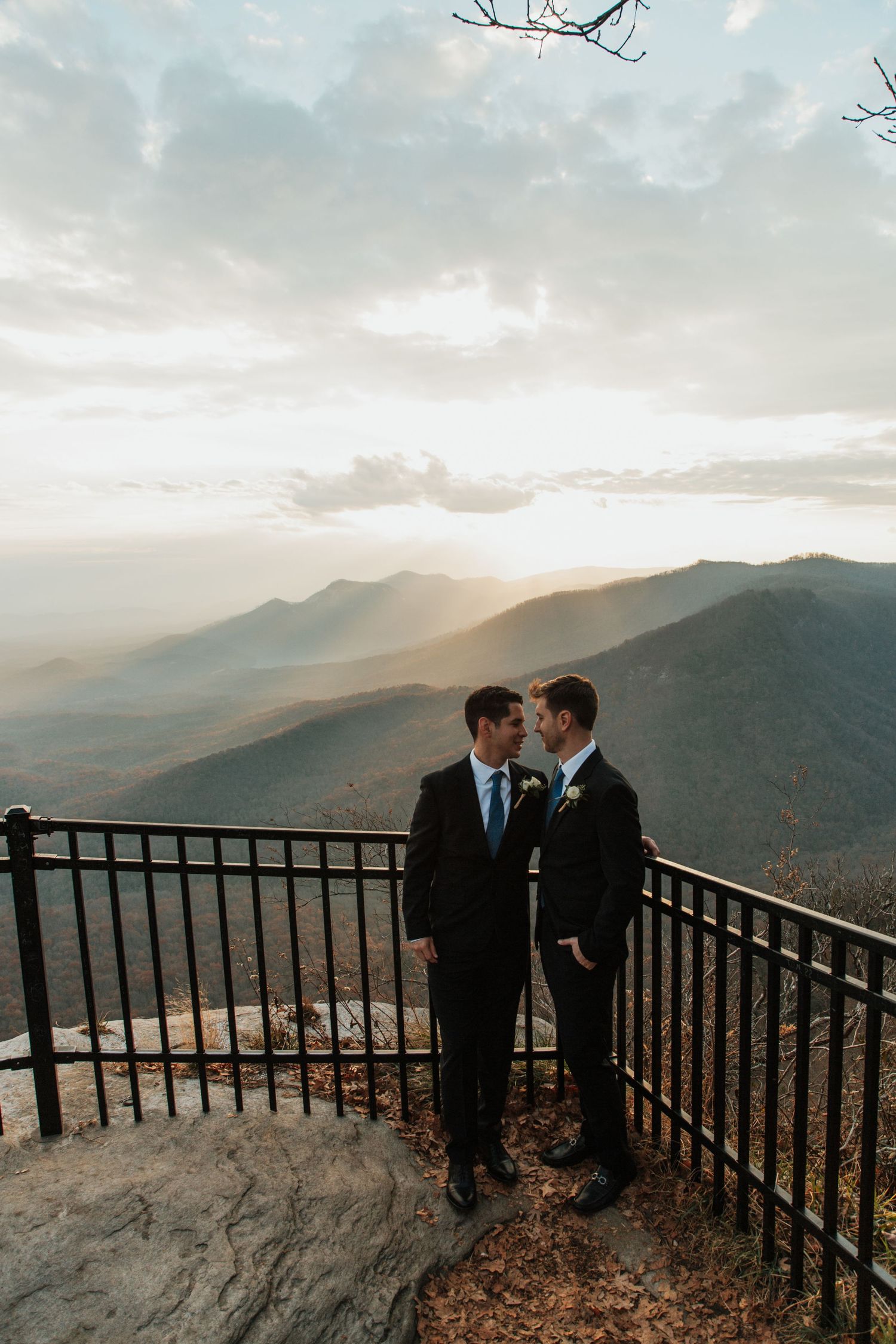 Two grooms in dark suits share an intimate moment on a scenic mountain overlook at golden hour sunset. caesars head state park