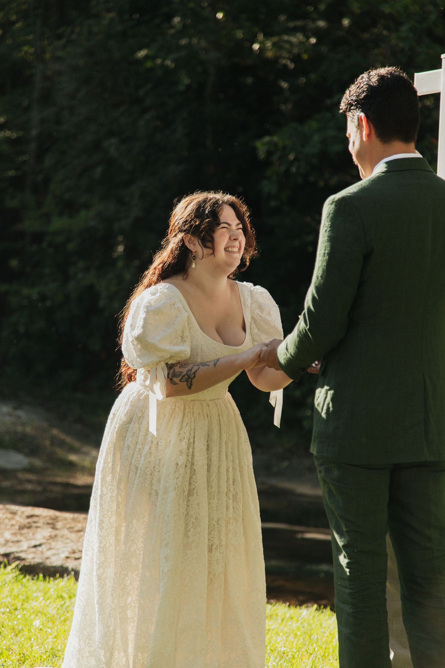 Smiling bride in white lace gown holding hands with groom in green suit during outdoor ceremony. venue at ballenger bridge landrum sc
