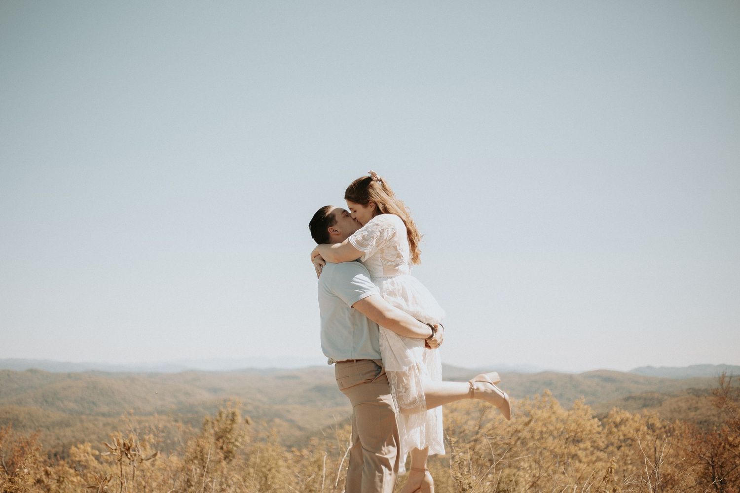 A man lifts a woman in a white dress as they kiss outdoors with scenic hills in the background.