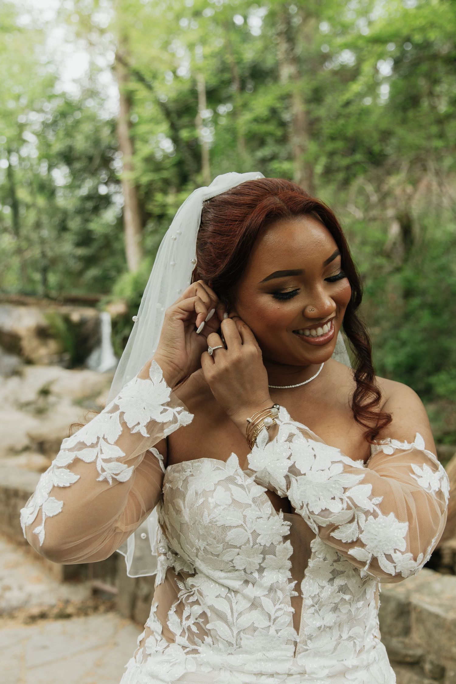 Smiling bride in lace off-shoulder wedding gown adjusting earring with veil in forest setting.  falls park greenville sc