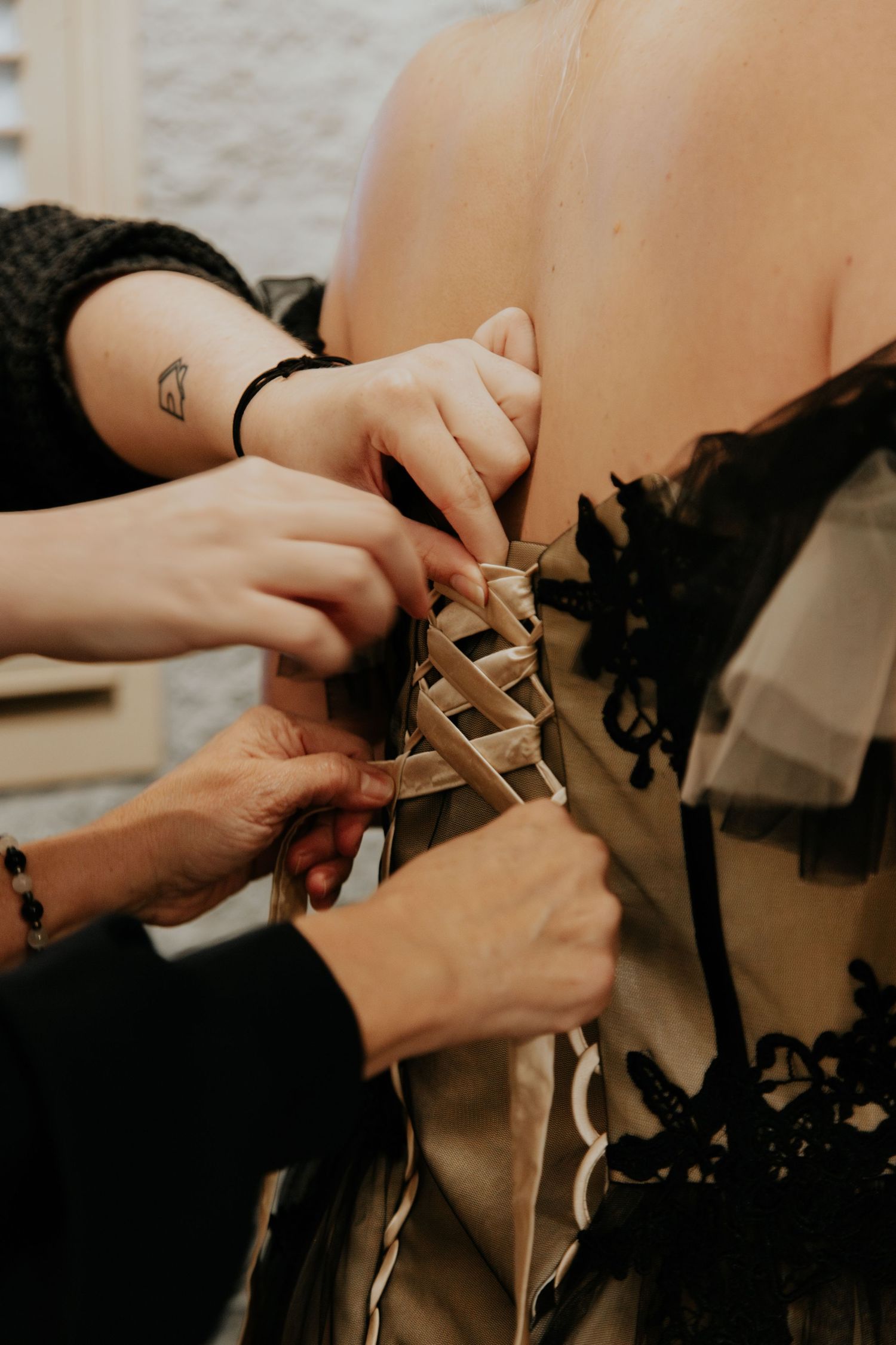 Two people lace up the corset back of a black and champagne wedding gown on a bride.