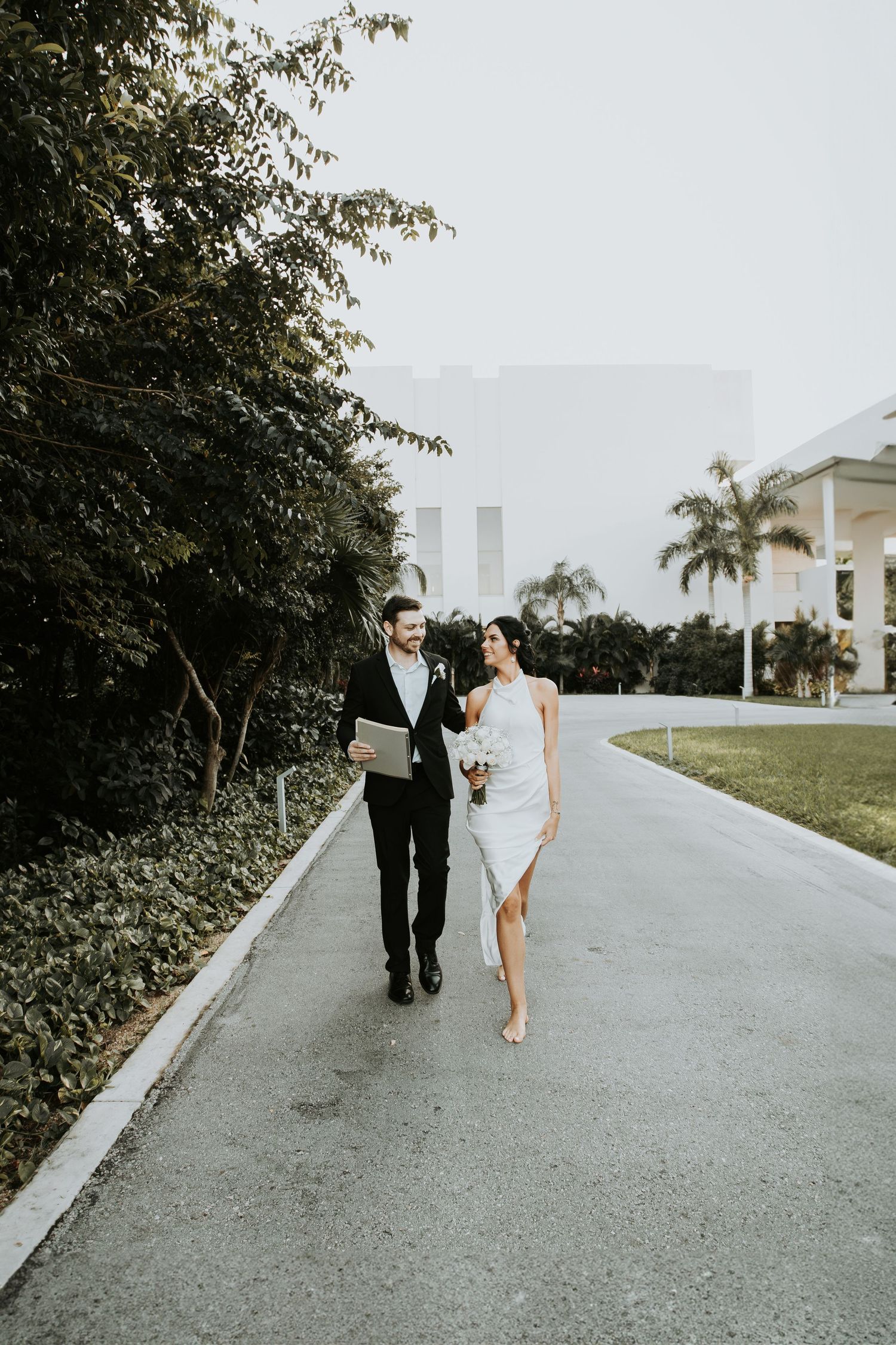Bride and groom walking hand in hand down a palm tree-lined path at outdoor venue. riviera maya cancun mexico