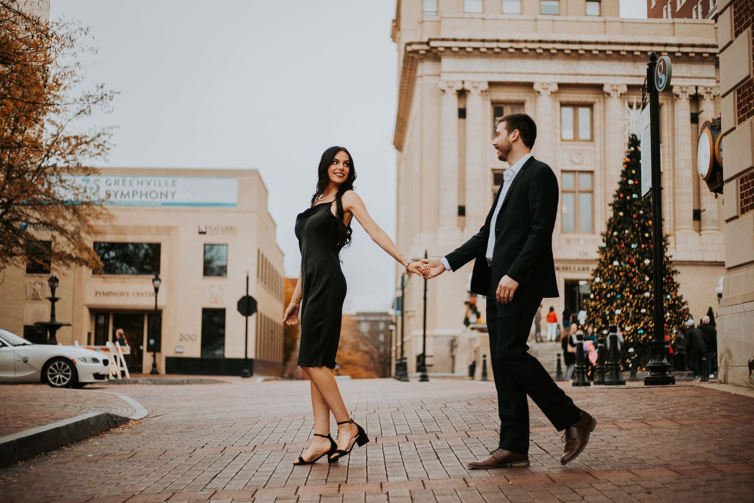 Couple holding hands and walking in a festive outdoor plaza with a Christmas tree. downtown main st greenville sc