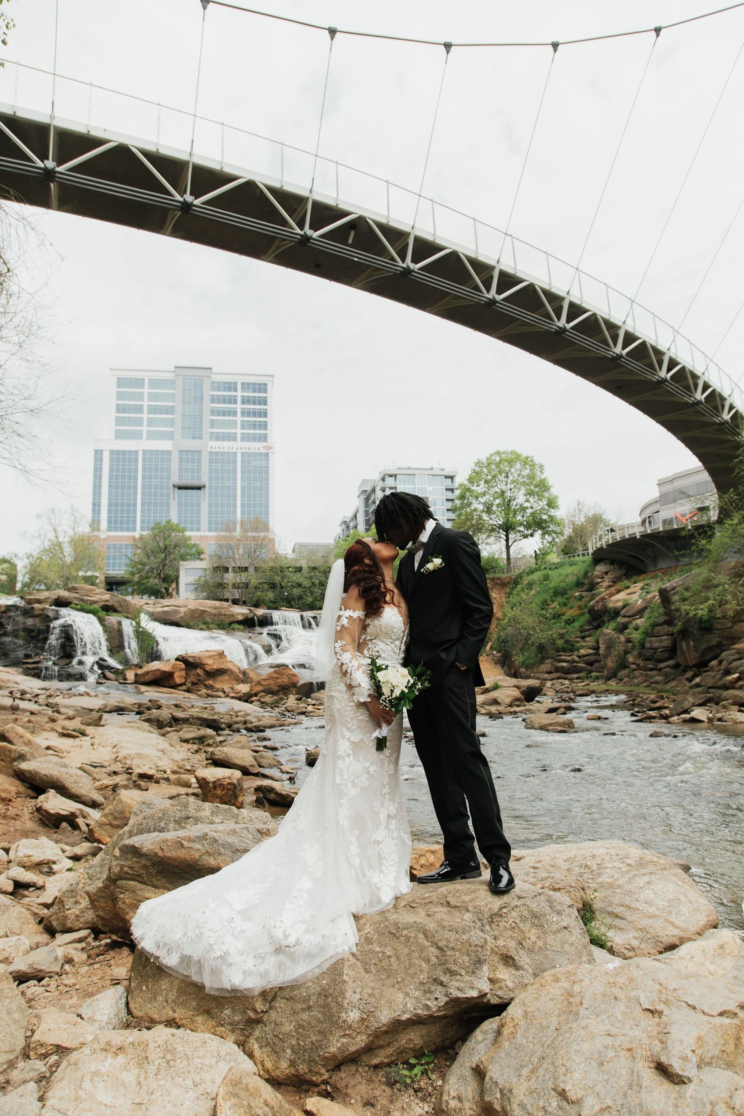 Bride and groom kissing on rocks beside a waterfall under an arched bridge with city buildings in the background.