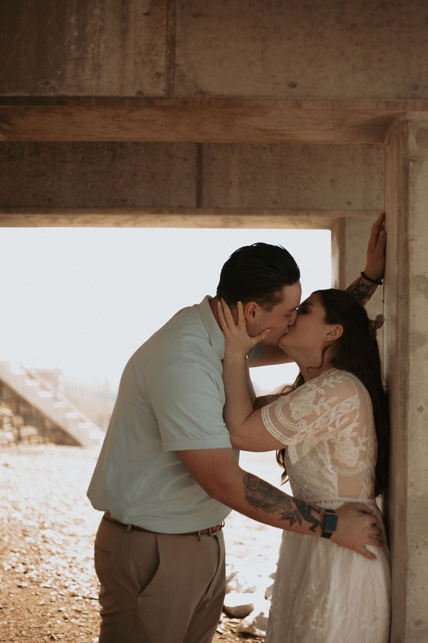 Couple sharing a romantic kiss against a concrete wall in an outdoor rustic setting, man in blue shirt, woman in white lace dress.