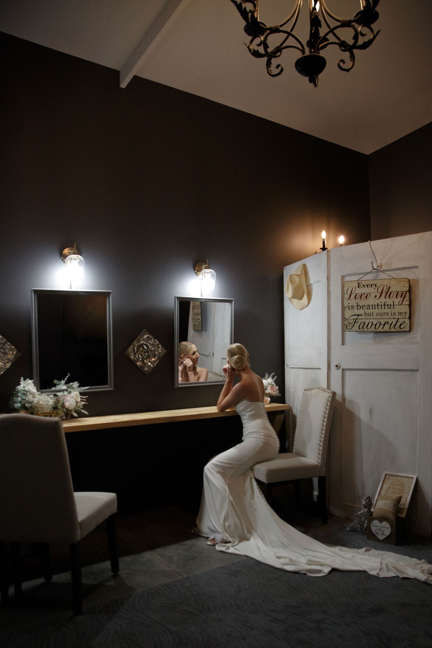 Bride in white gown sits at vanity mirror in dimly lit bridal suite, preparing for her wedding day.