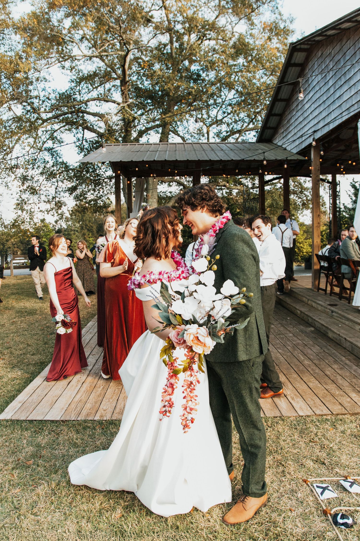 Bride and groom share a kiss outdoors at rustic wedding venue, surrounded by bridesmaids in red dresses.
