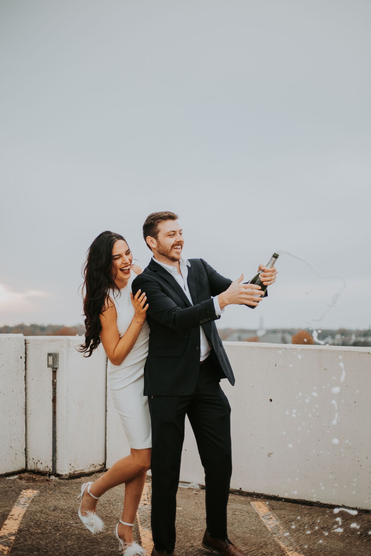 Joyful couple popping champagne bottle on rooftop, woman in white dress and man in black suit celebrating engagement. greenville sc