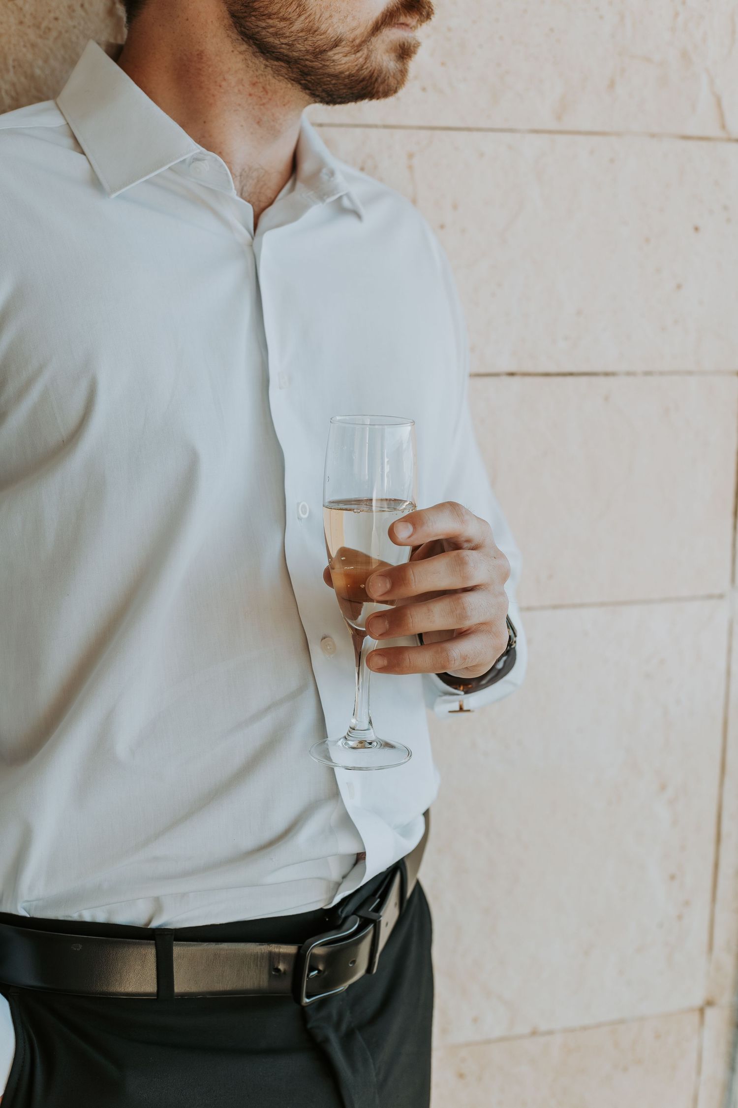 Groom in white dress shirt holds champagne flute against beige stone wall. - riviera maya resort (cancun, Mexico)