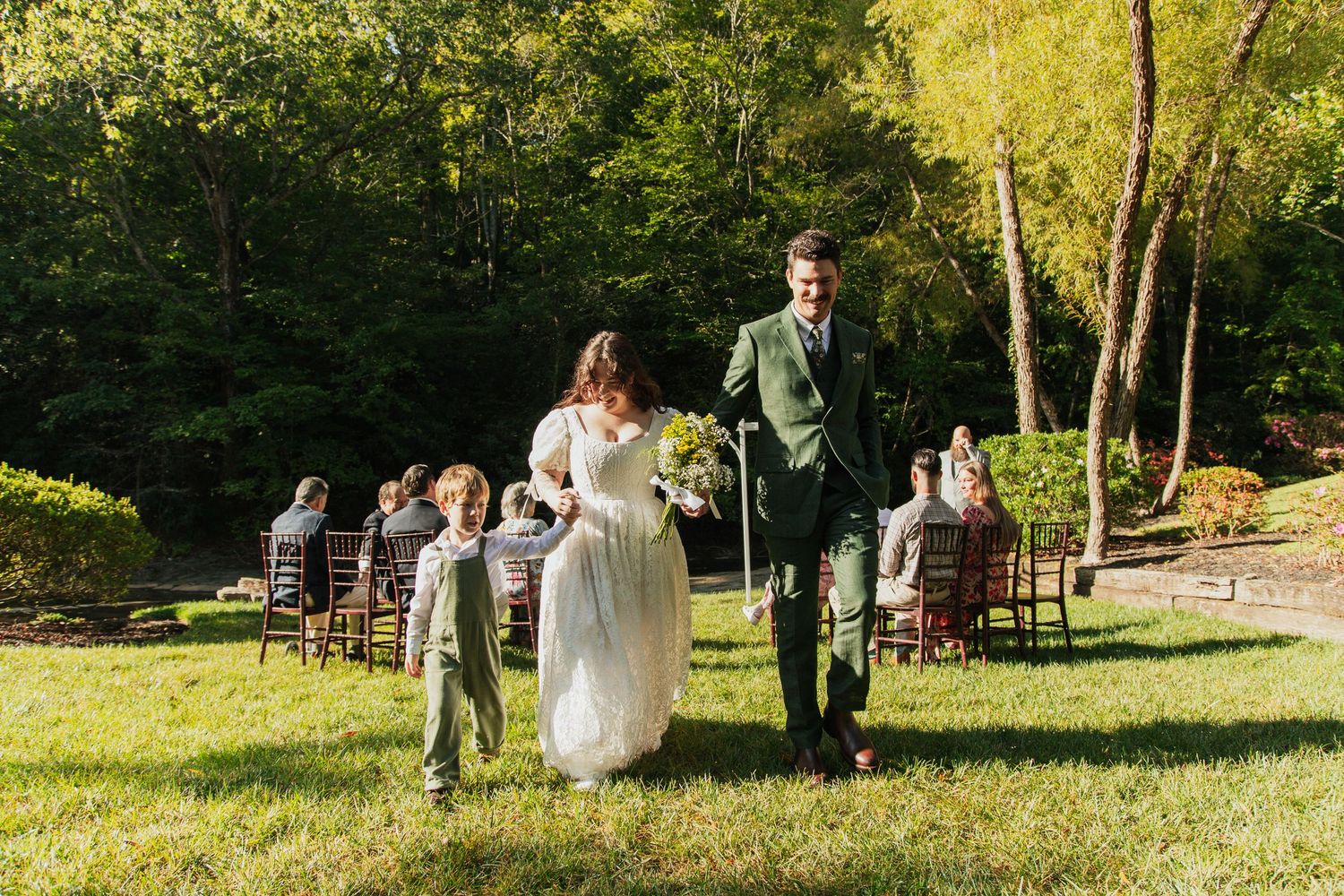 Newlyweds joyfully walking down grassy aisle after outdoor woodland wedding ceremony with guests seated. venue at ballenger bridge landrum s