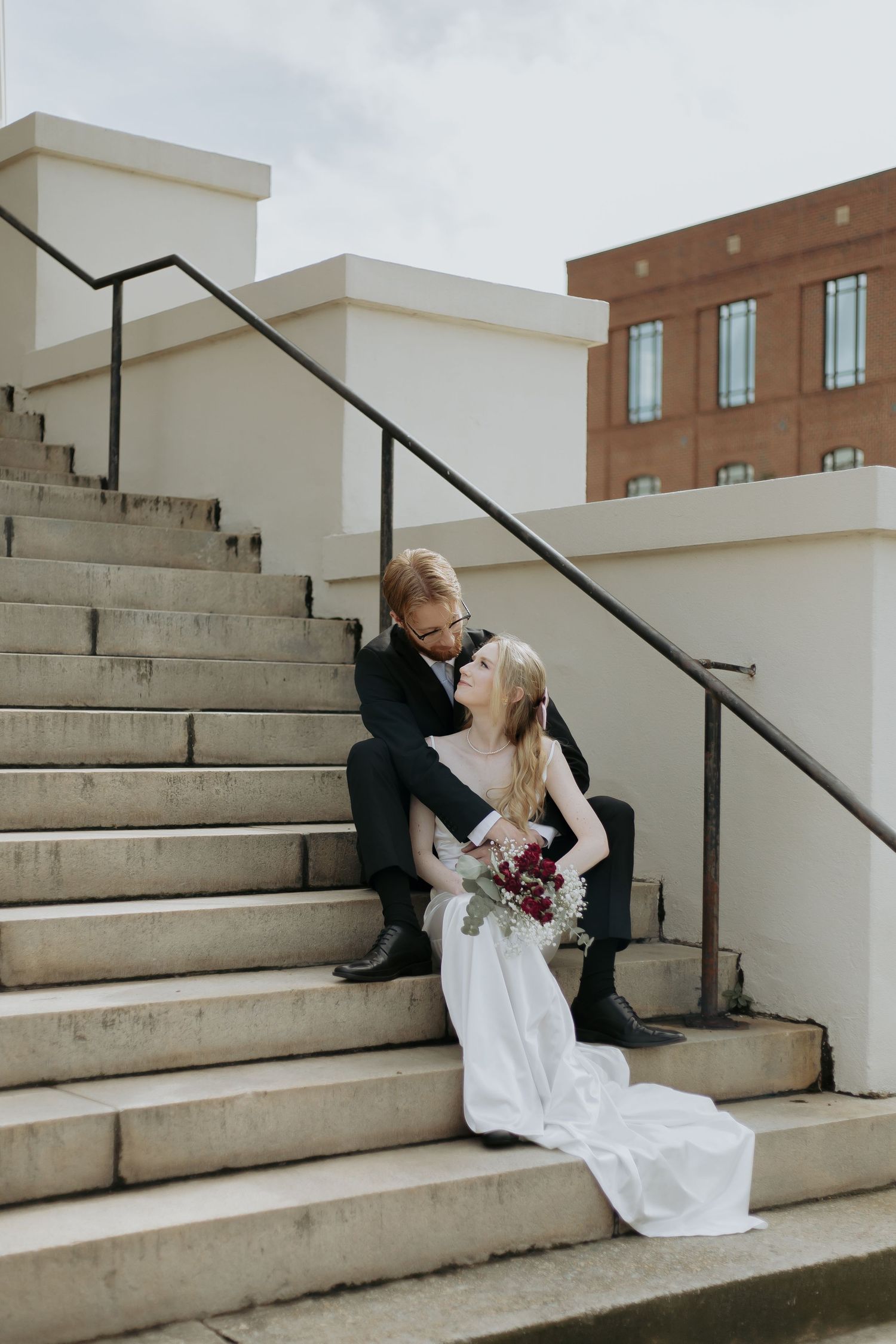 Bride and groom sitting on outdoor stone steps, she holds a burgundy bouquet in a white gown, he wears a black suit.