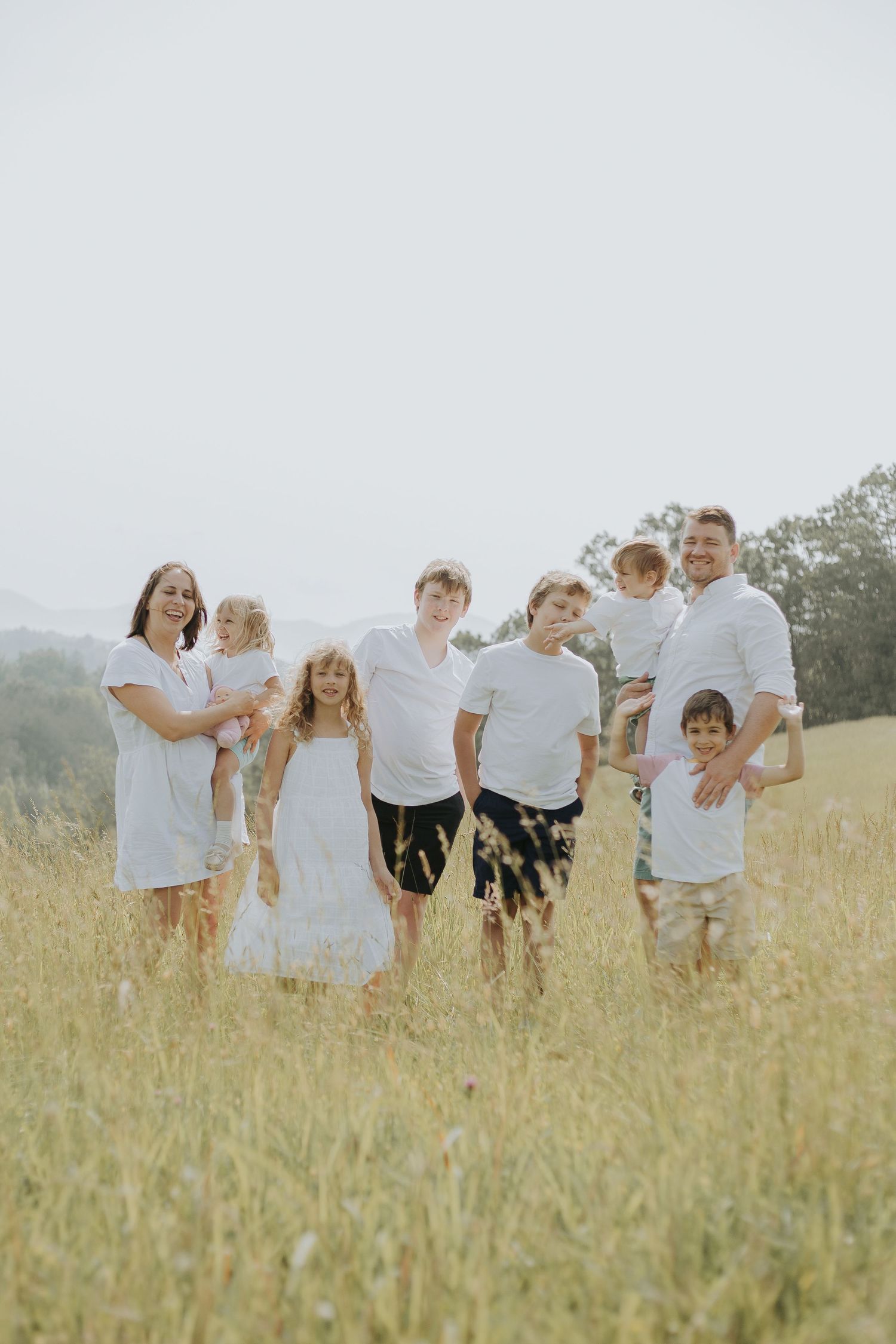 Large family of eight posing together in a golden meadow field, wearing white outfits on a bright sunny day.
