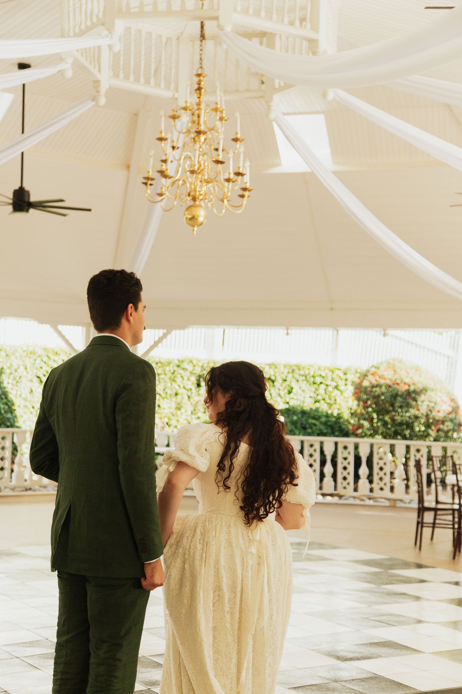 Bride and groom standing hand in hand beneath gold chandelier in elegant white draped wedding venue. venue at ballenger bridge landrum sc