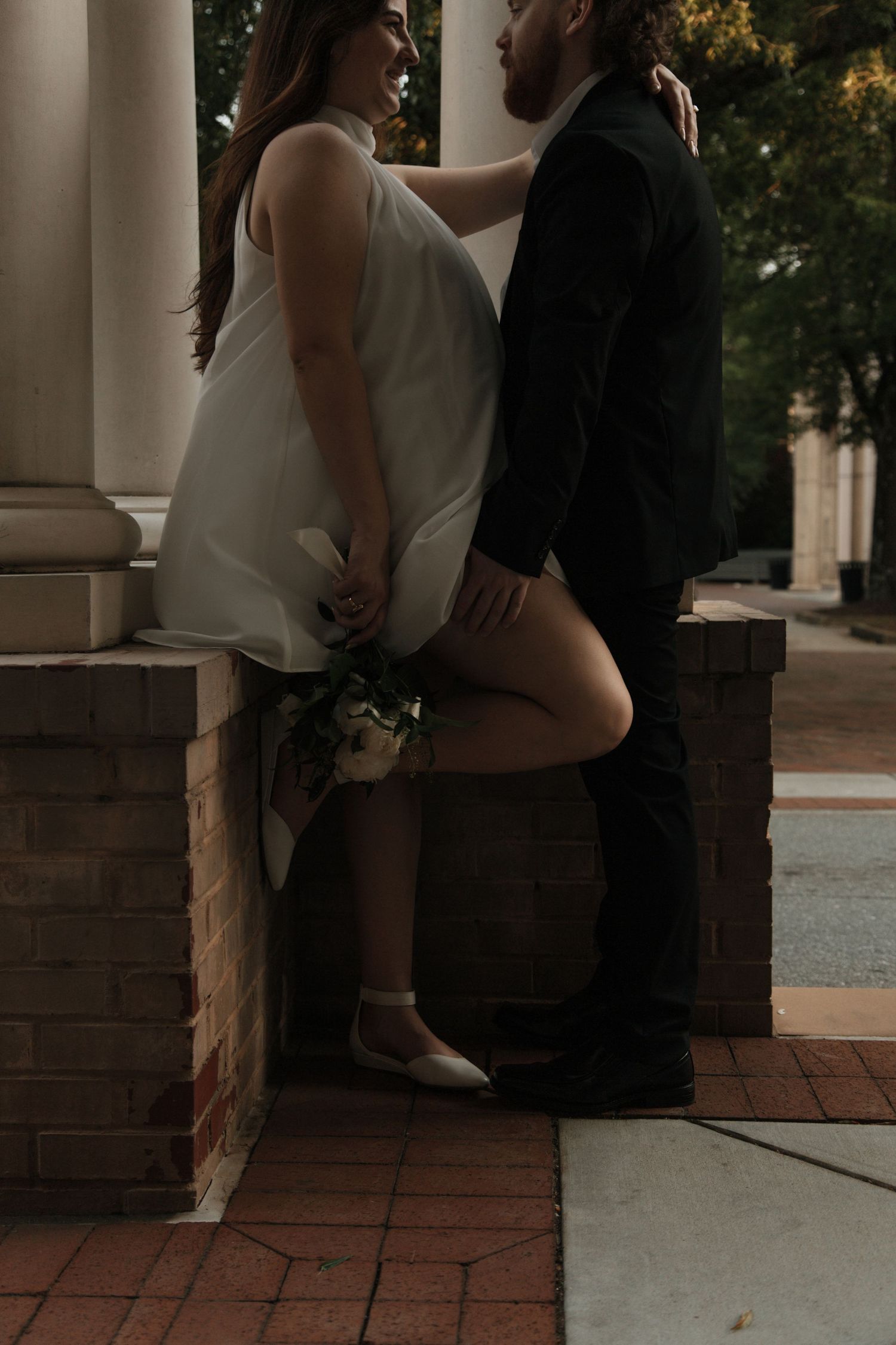 Bride in white mini dress leaning against brick column with groom in intimate wedding portrait. anderson, sc