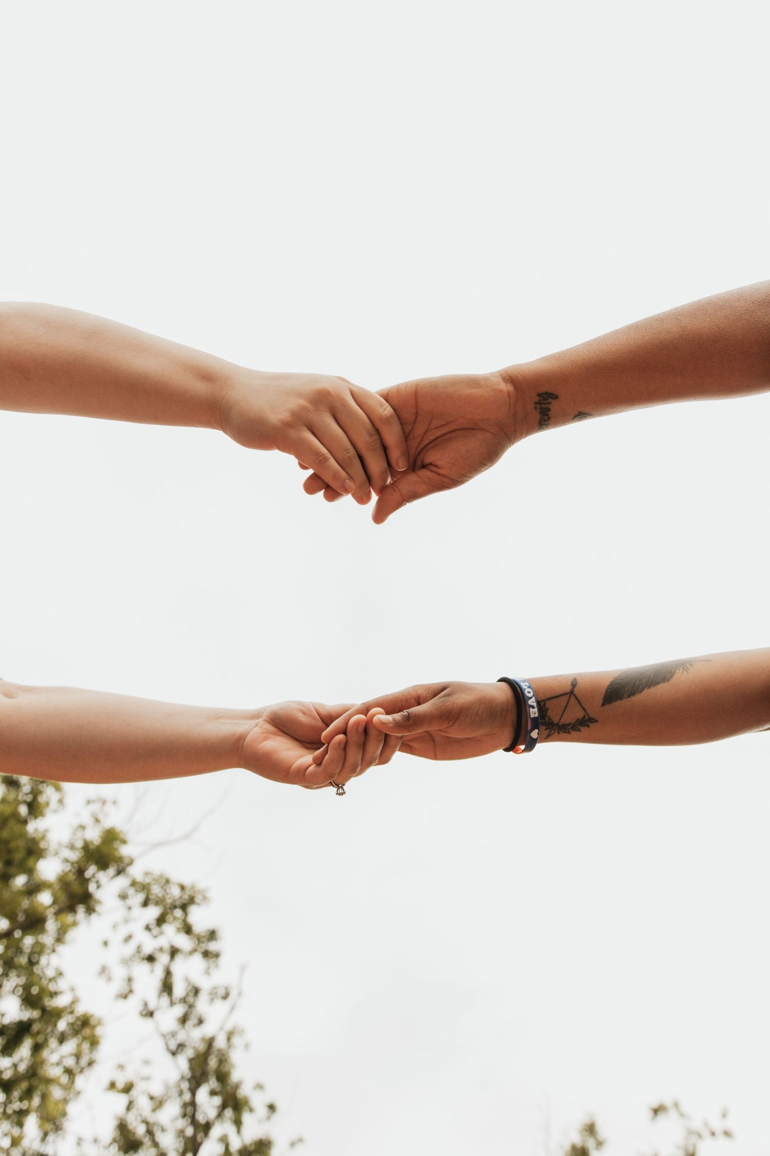 Two people holding hands with tattoos and a bracelet visible against a bright sky with trees in the background.