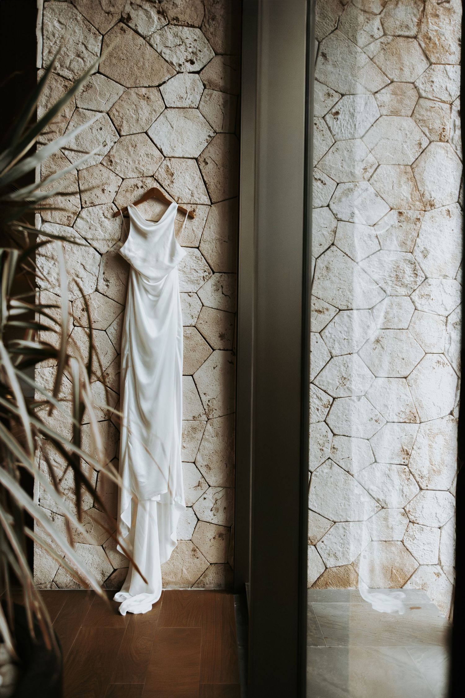 Elegant white satin wedding dress hanging on wooden hanger against stone wall. - riviera maya resort (cancun, Mexico)