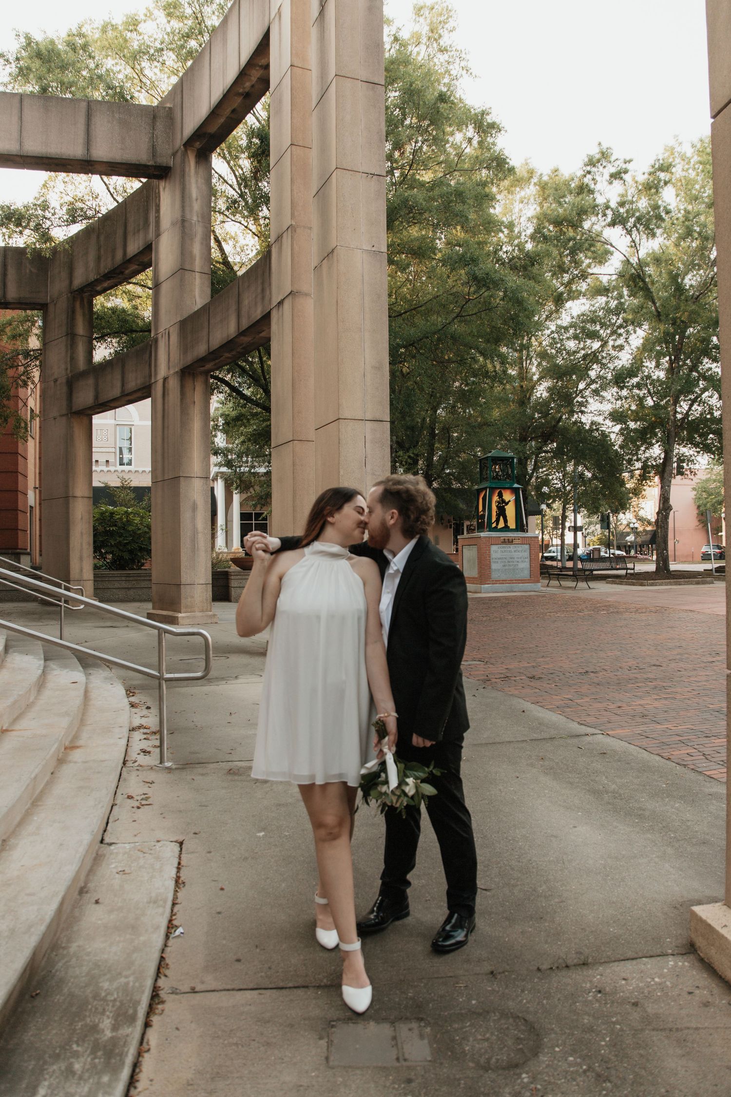 Bride and groom sharing a kiss on urban plaza walkway surrounded by stone columns and trees. downtown anderson court house south carolina