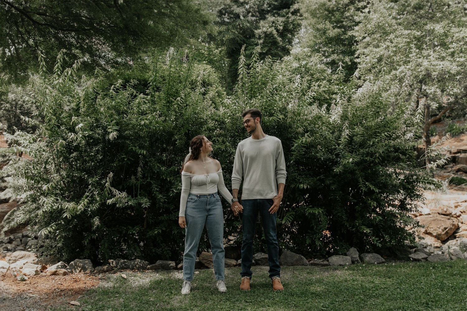 Couple holding hands smiling at each other in a lush green park setting, wearing casual outfits.