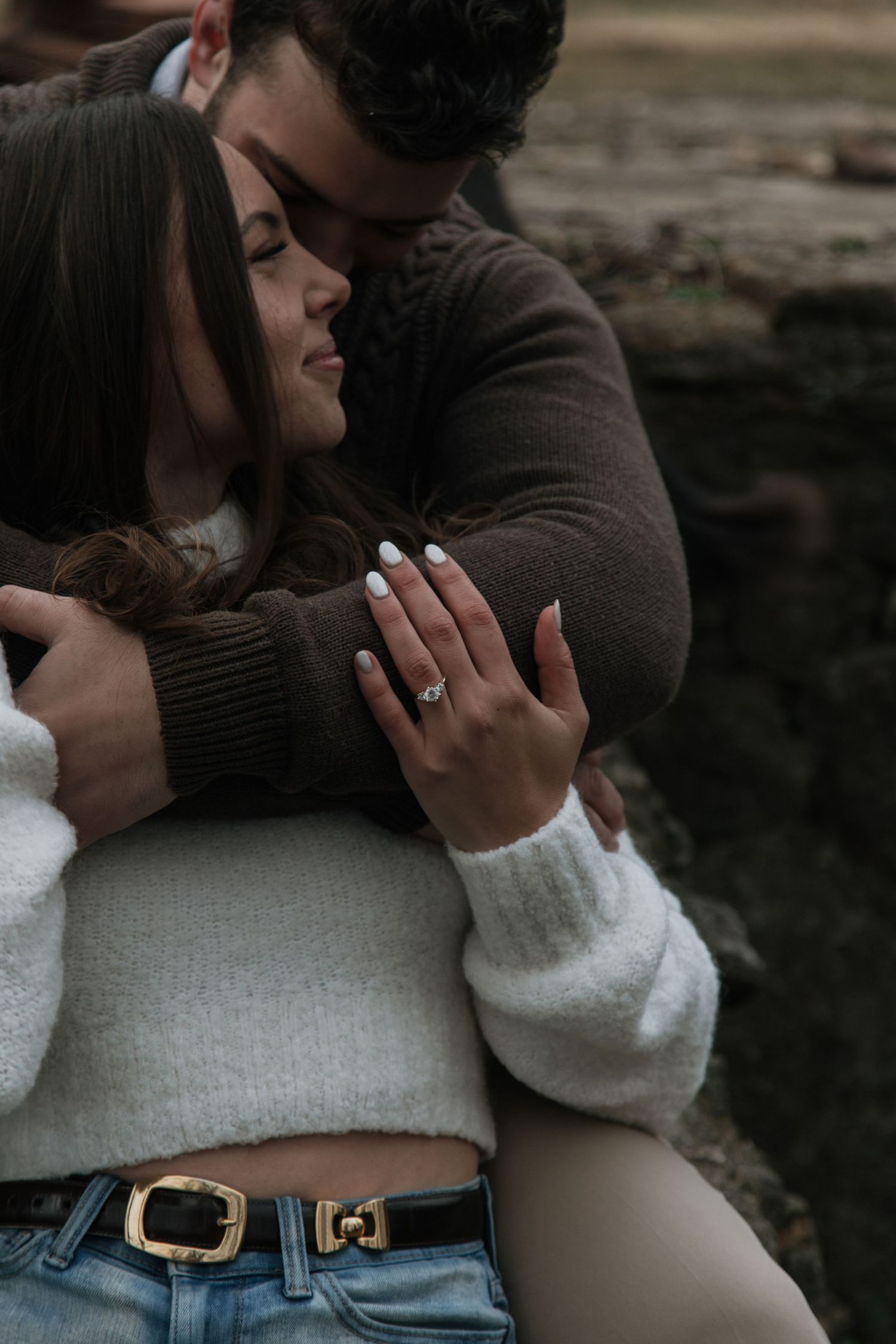 Couple embracing outdoors, woman displaying engagement ring on hand, wearing white sweater and jeans.